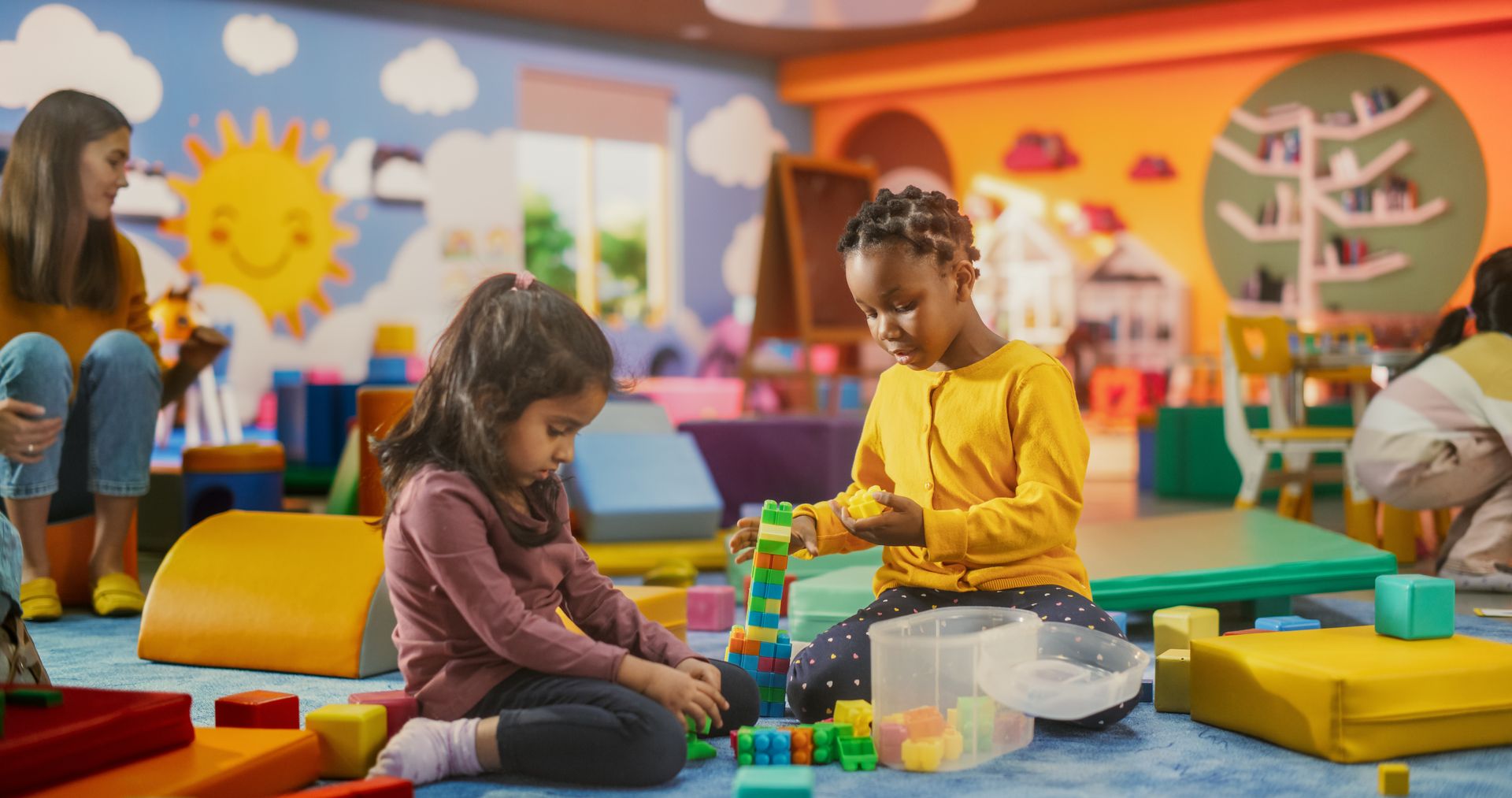 Two children playing with colorful blocks in a playroom, a teacher supervises.