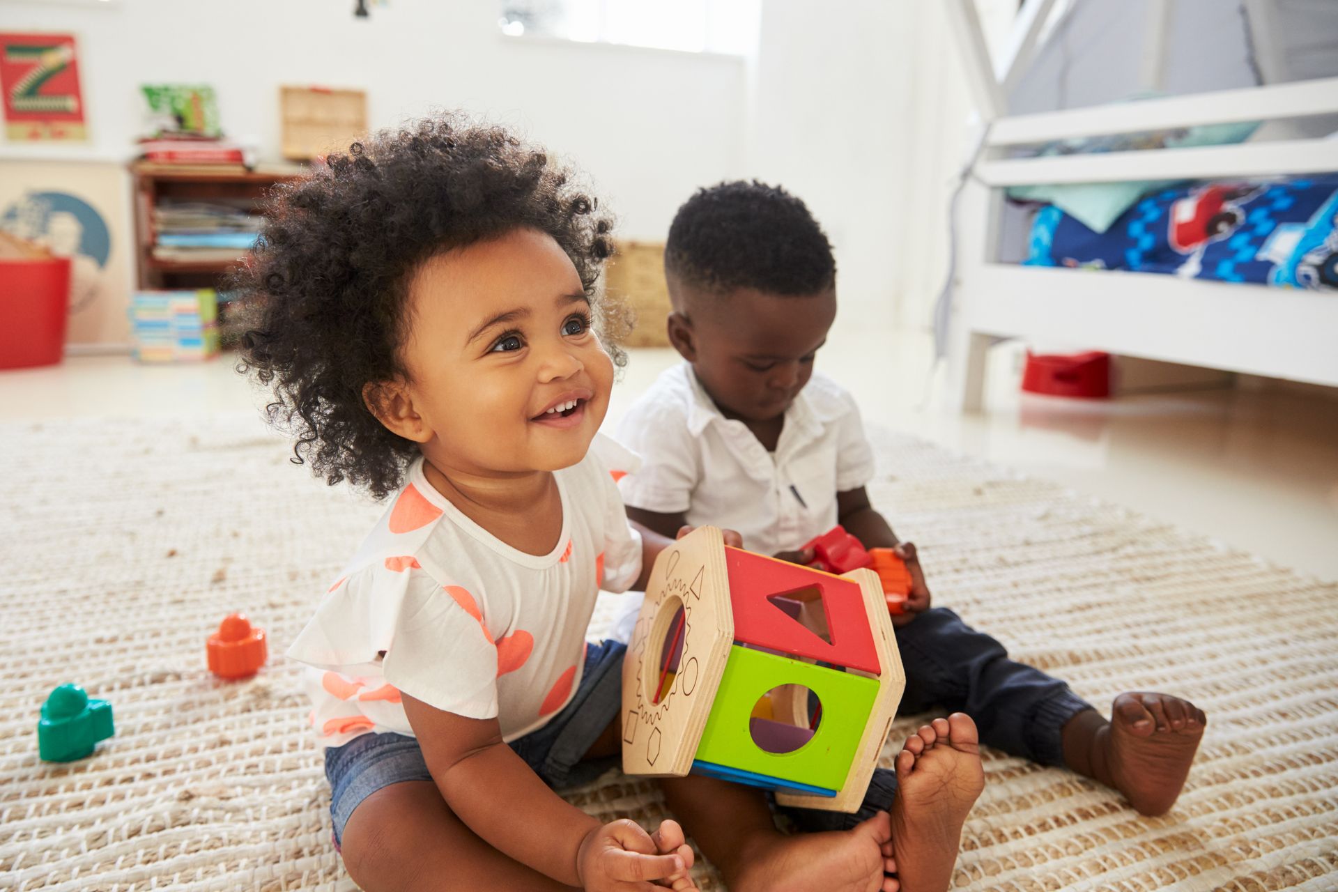 Two toddlers playing with a shape sorter on a rug in a brightly lit room.