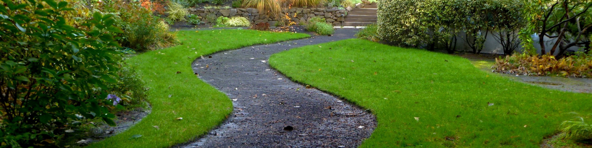 A winding, dark gravel path cuts through a vibrant green lawn in a landscaped garden with surrounding bushes.