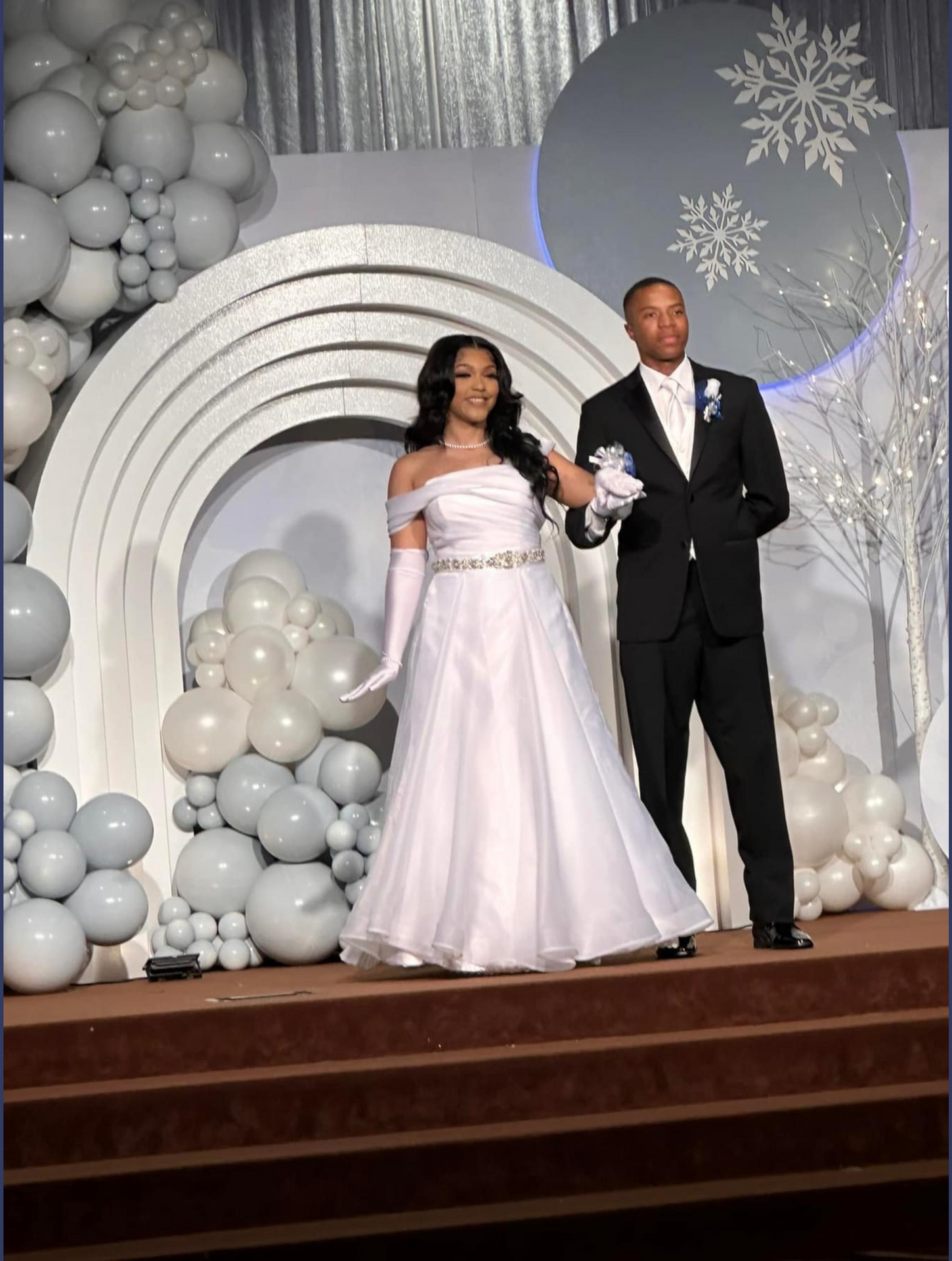 A debutant and beau are standing on a stage with balloons in the background