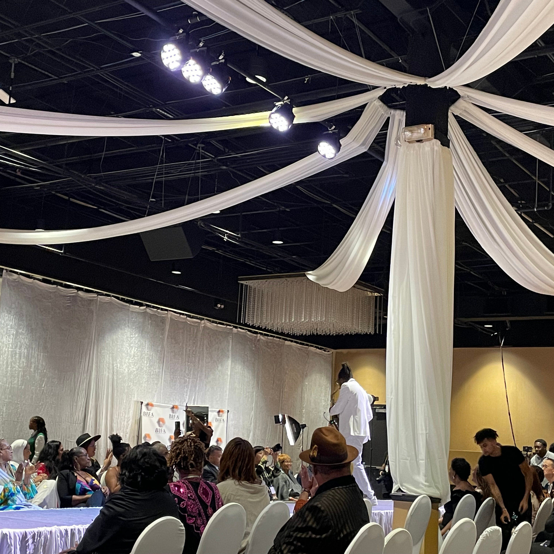 A group of people are sitting in a large room with white curtains hanging from the ceiling