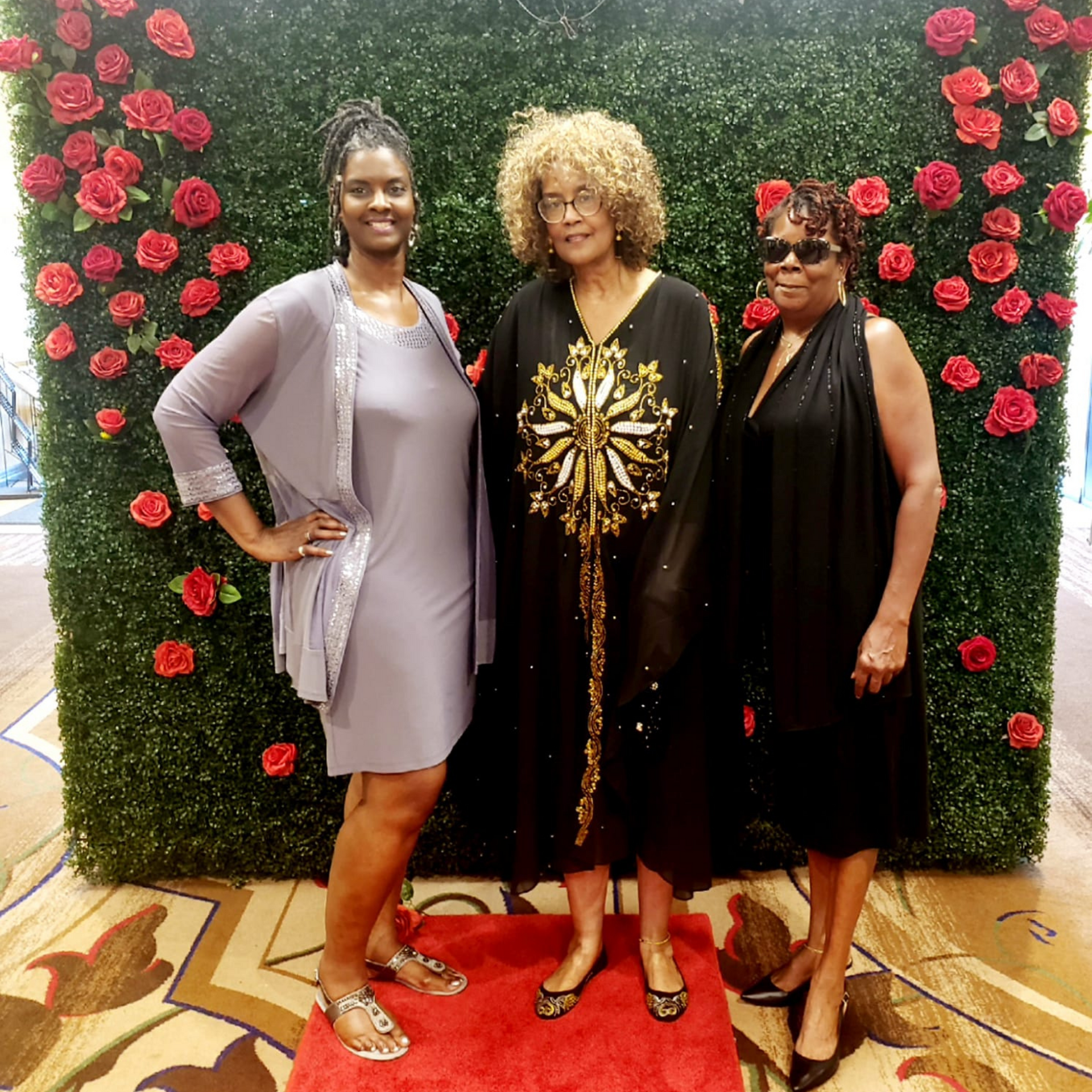 Three women are posing for a picture in front of a wall of roses.