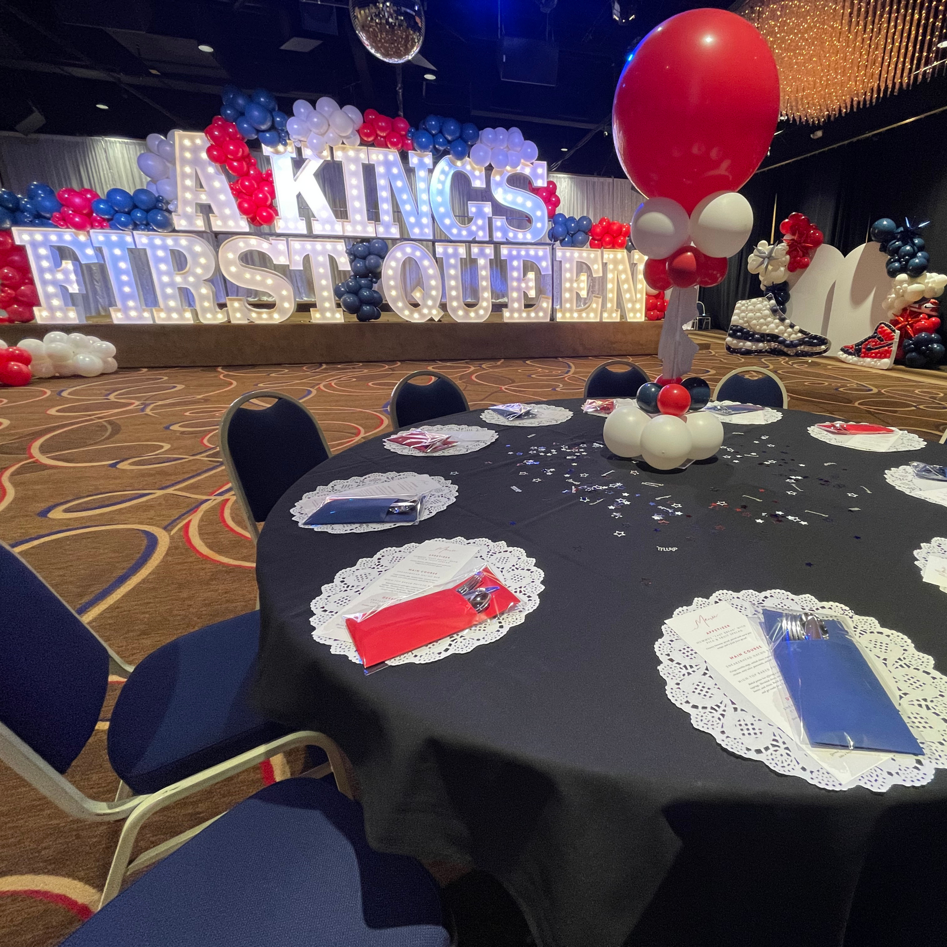A kings first queen sign is behind a table with balloons on it