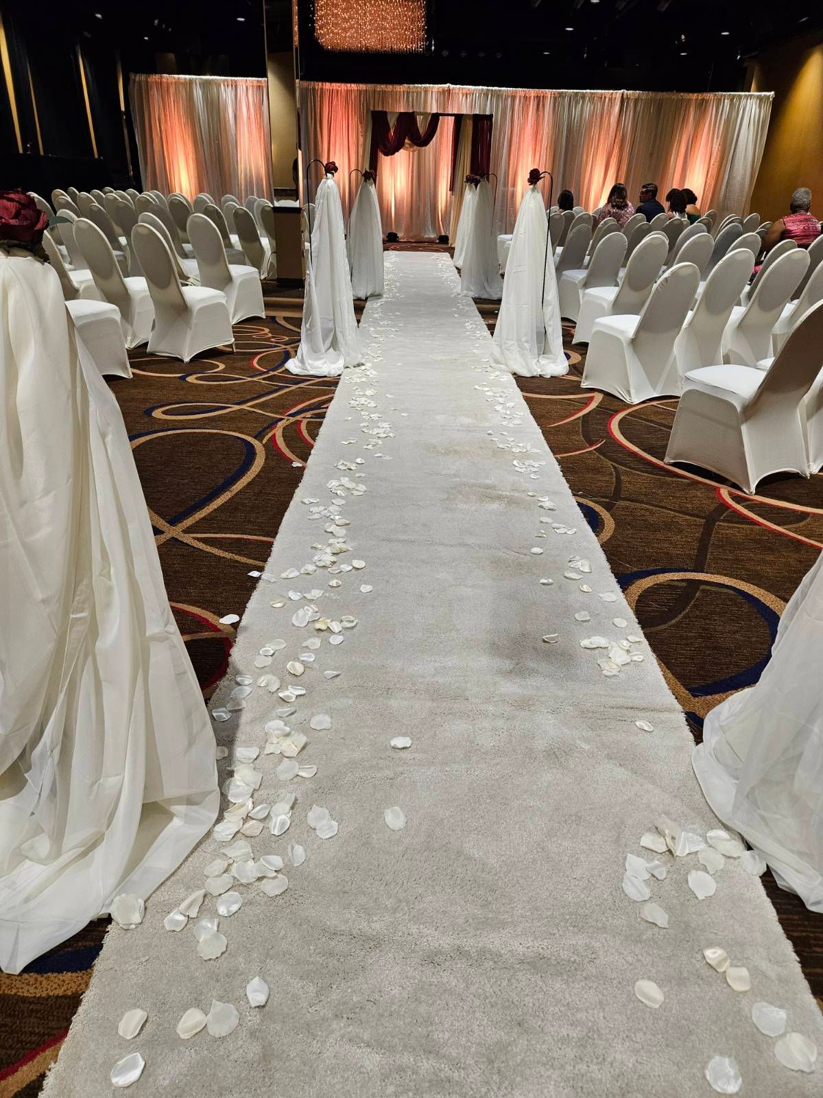 A wedding ceremony is taking place in a large room with white chairs and petals on the floor.