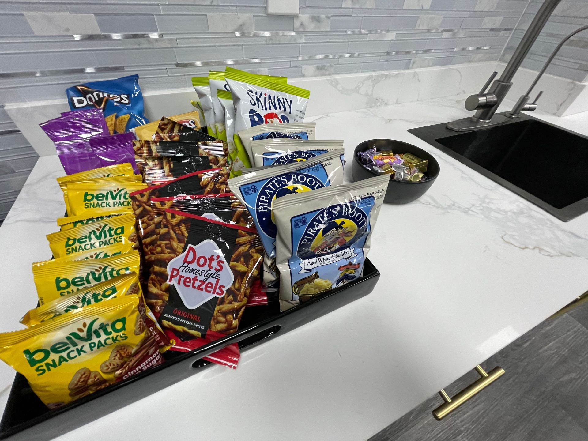 Snack tray on a white countertop with various chips, pretzels, and granola bars near a kitchen sink.