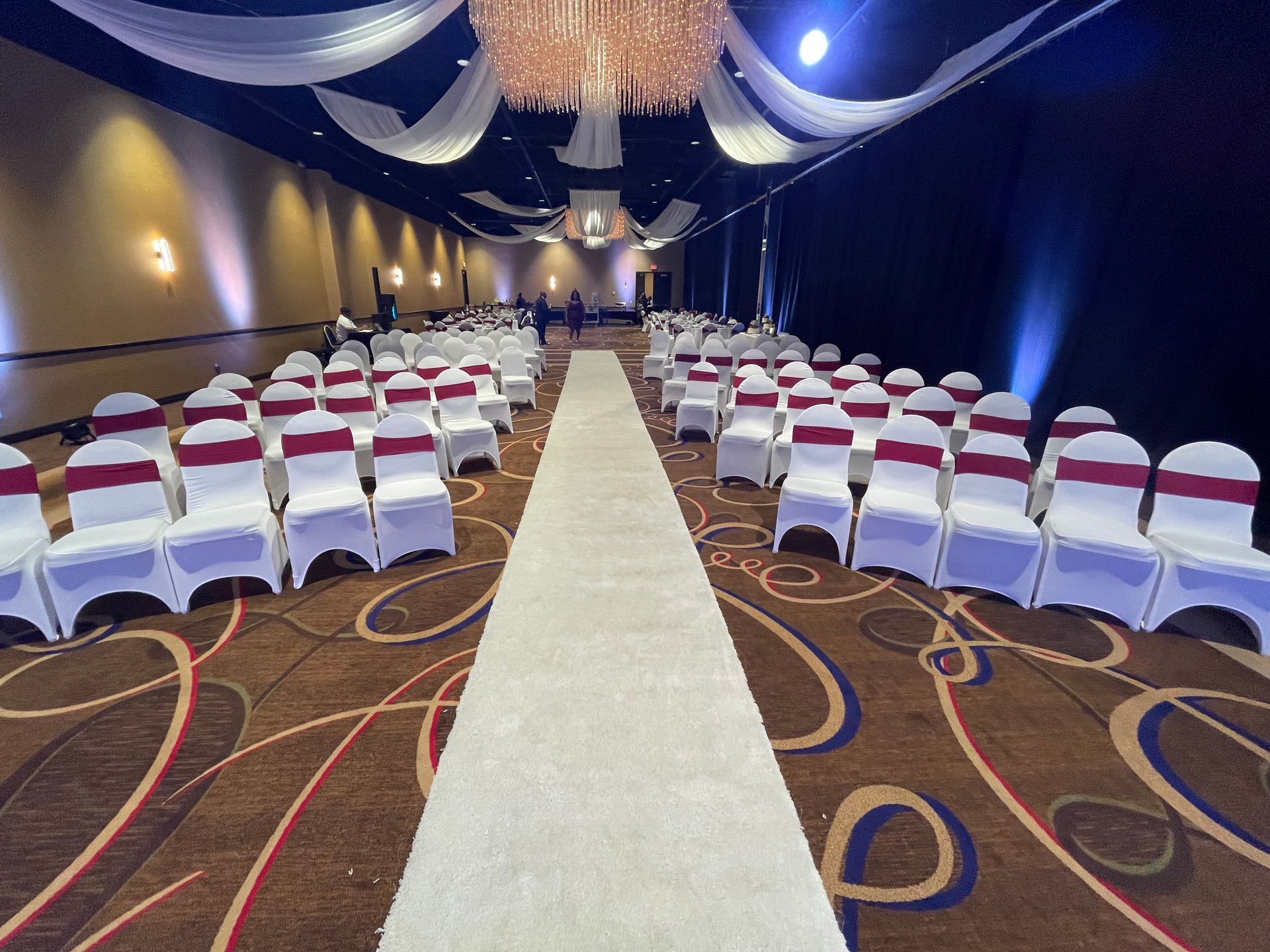 A long aisle in a room with white chairs and red sashes.