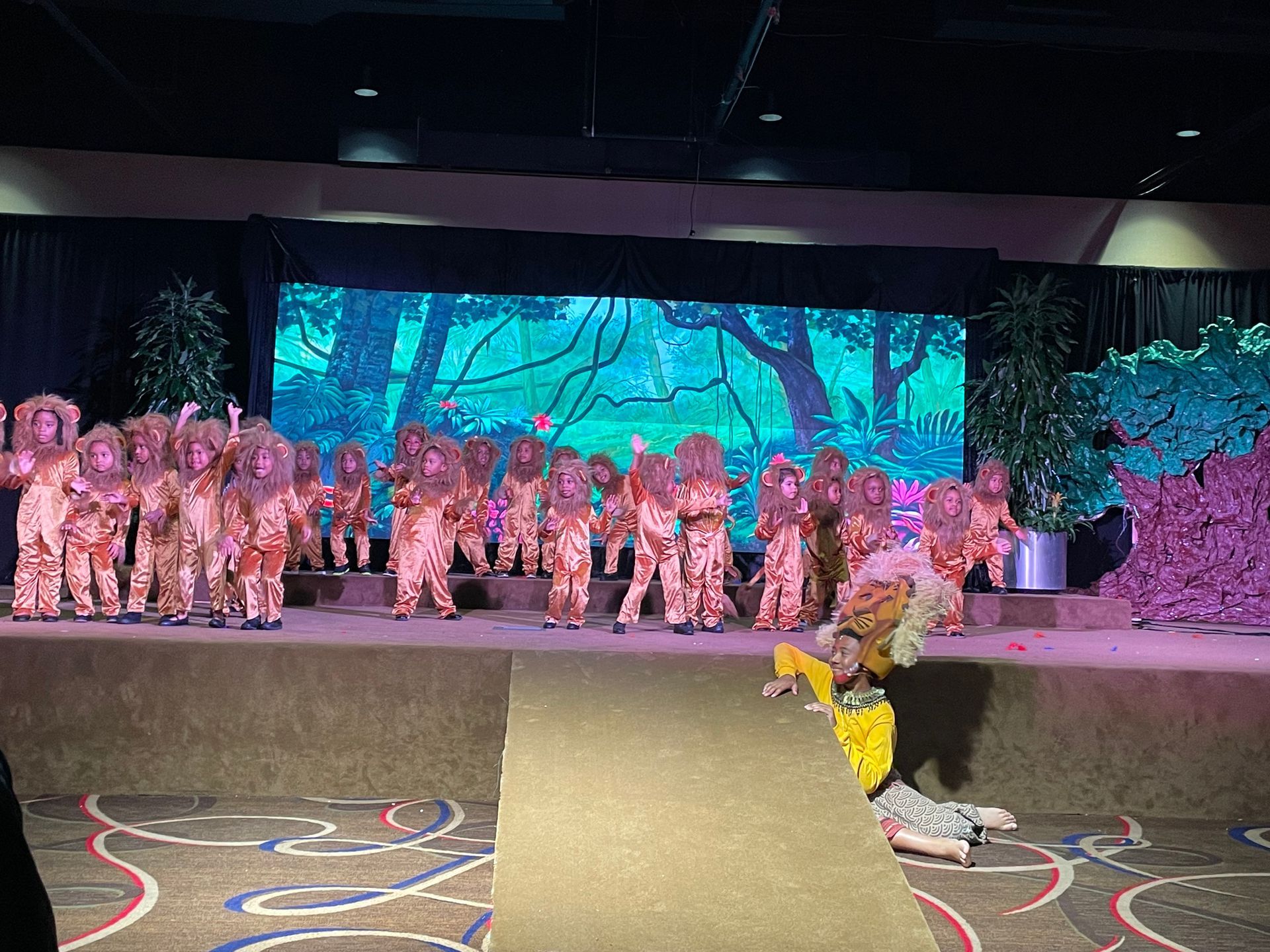 A group of children are dancing on a stage in front of a large screen.