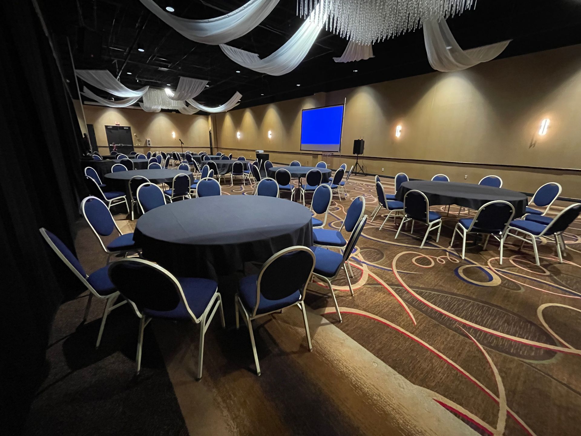 Large event space with round tables covered in black linens and blue chairs. A screen is visible.