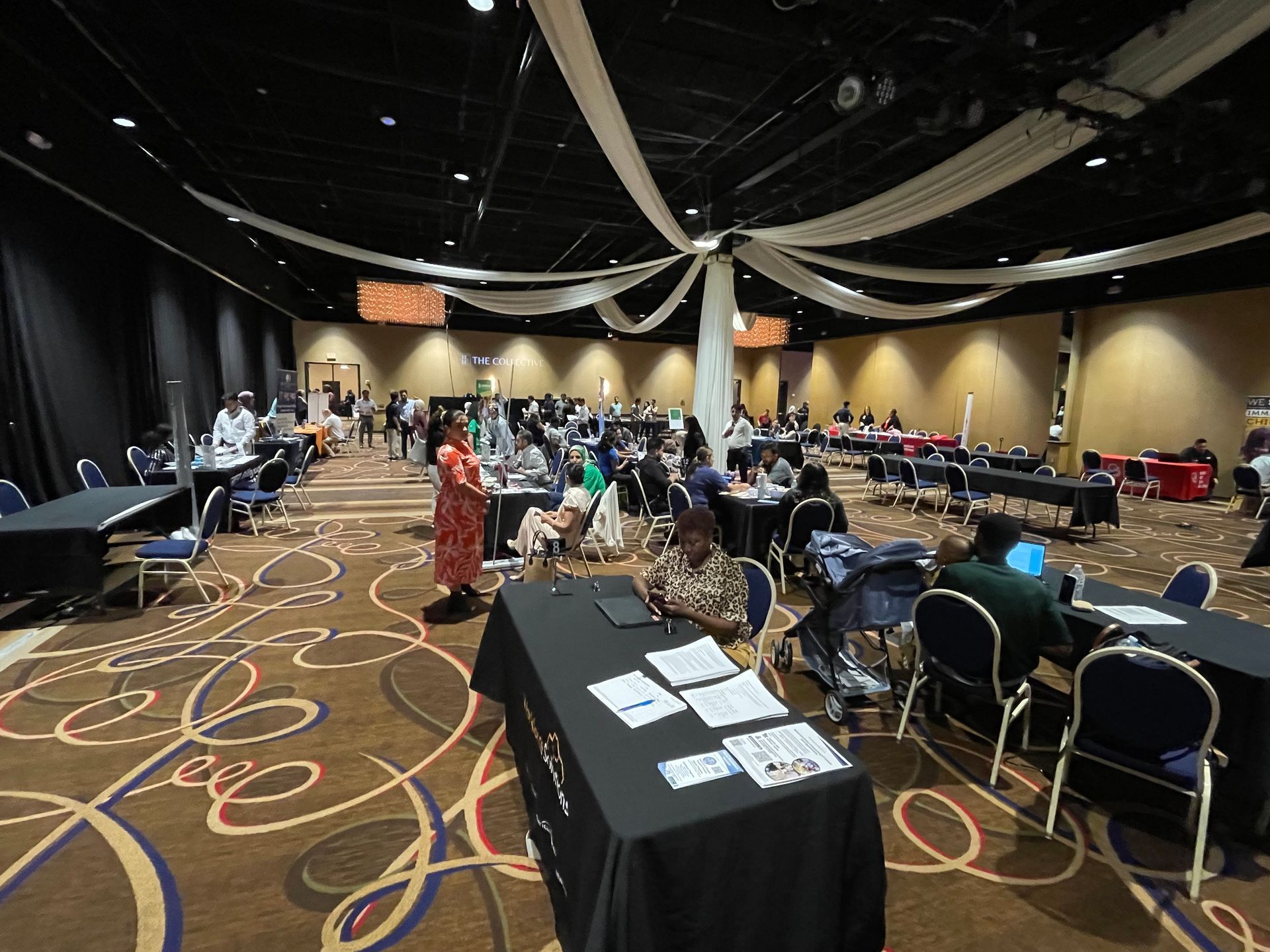 Large event space with tables, chairs, and people. A woman in a red patterned dress stands in the center.