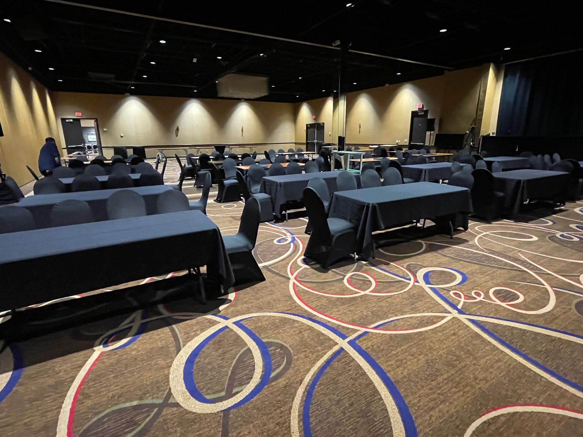 Empty event space with rows of tables covered in black cloth and chairs on patterned carpet.