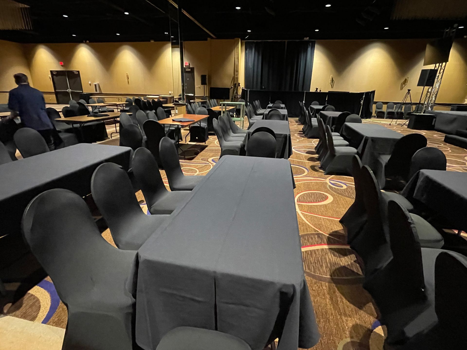 Empty event room with tables covered in black cloth and chairs, some people.