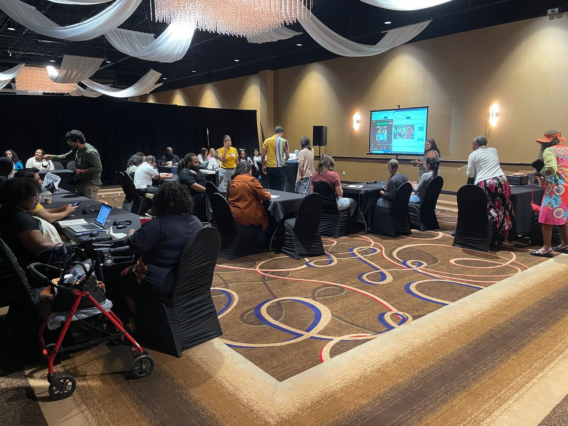 People at tables in a large event room; projector screen in the back, patterned carpet, decorative ceiling.