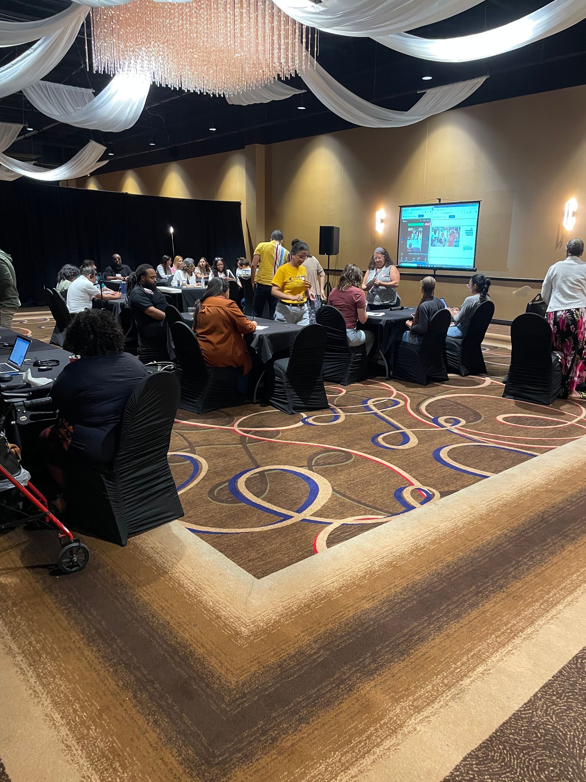 People seated at round tables in a conference hall, listening to a presentation on a screen.