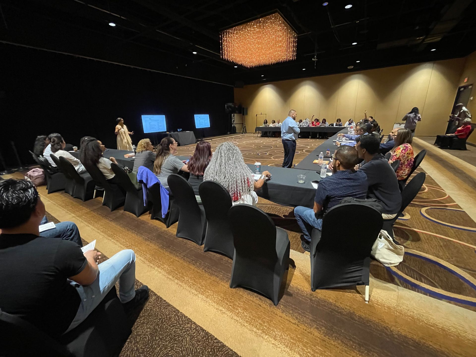 Audience in a conference room listening to a presenter, with screens in the background.