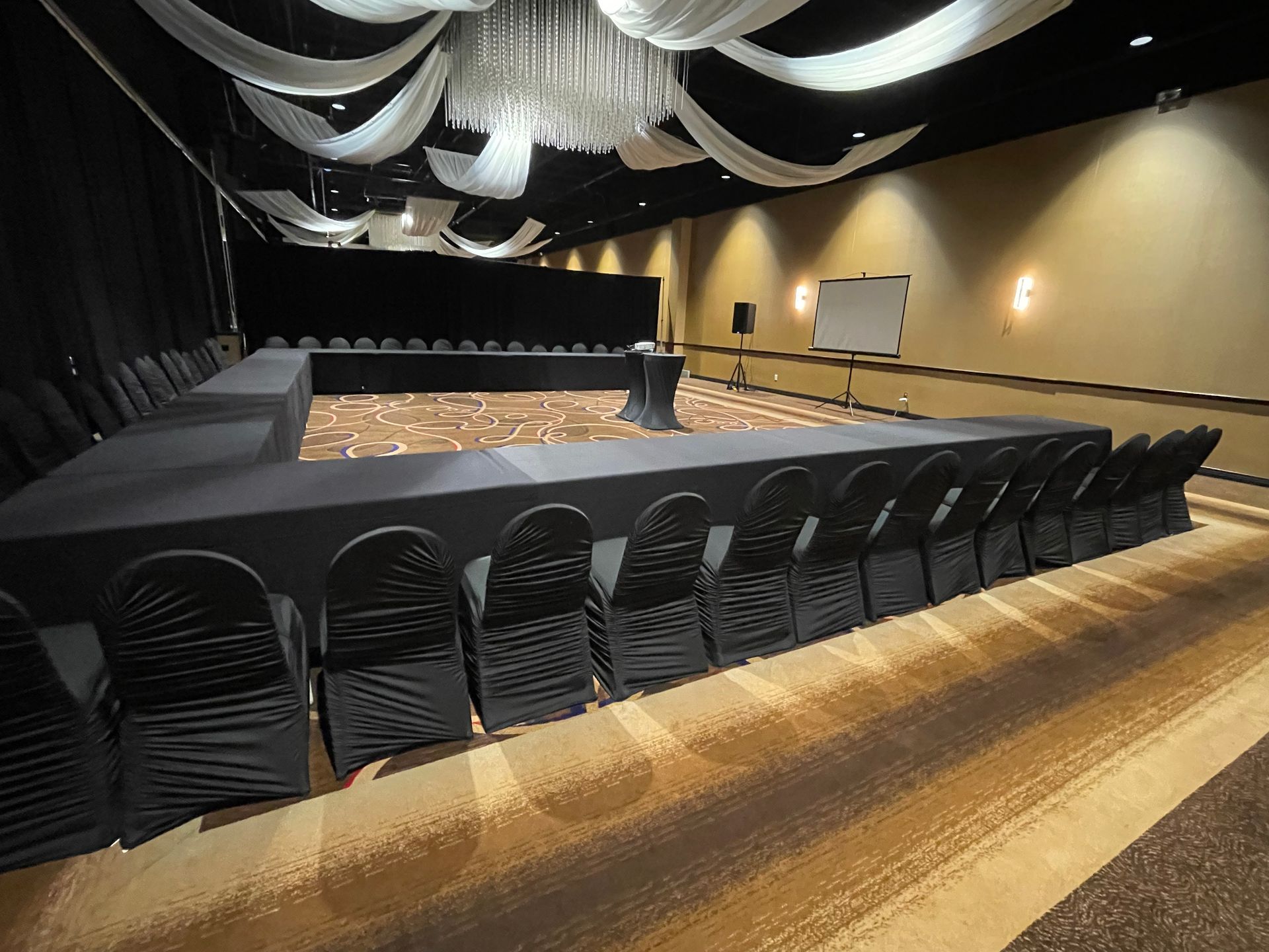 A U-shaped conference room with black chairs and a stage, draped ceiling, and projector screen.