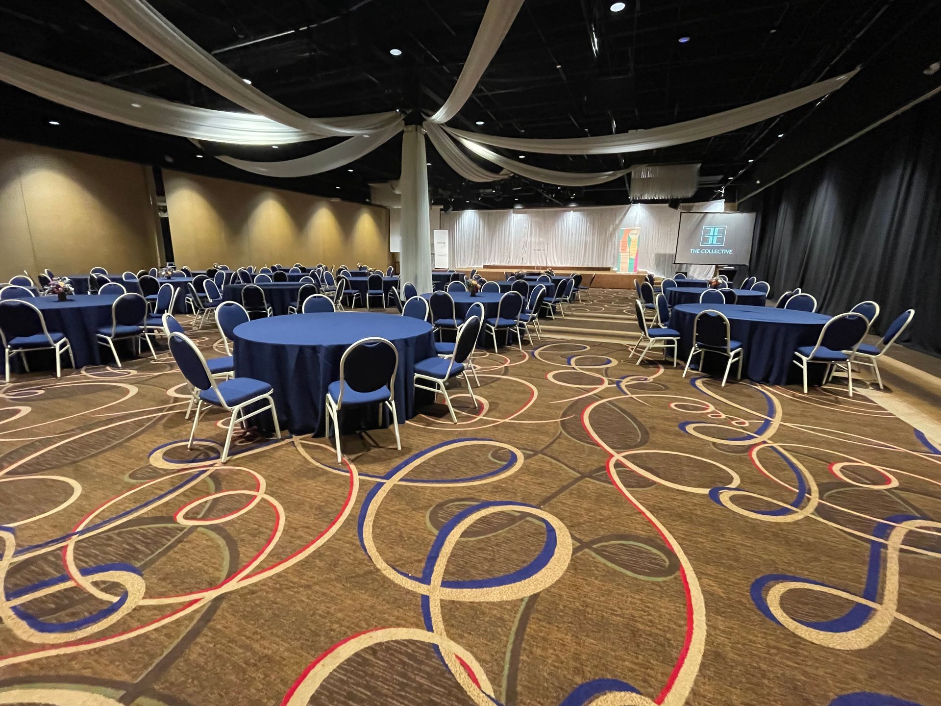 Large event space with round tables covered in blue cloths. Patterned carpet, stage, and decorative ceiling accents.