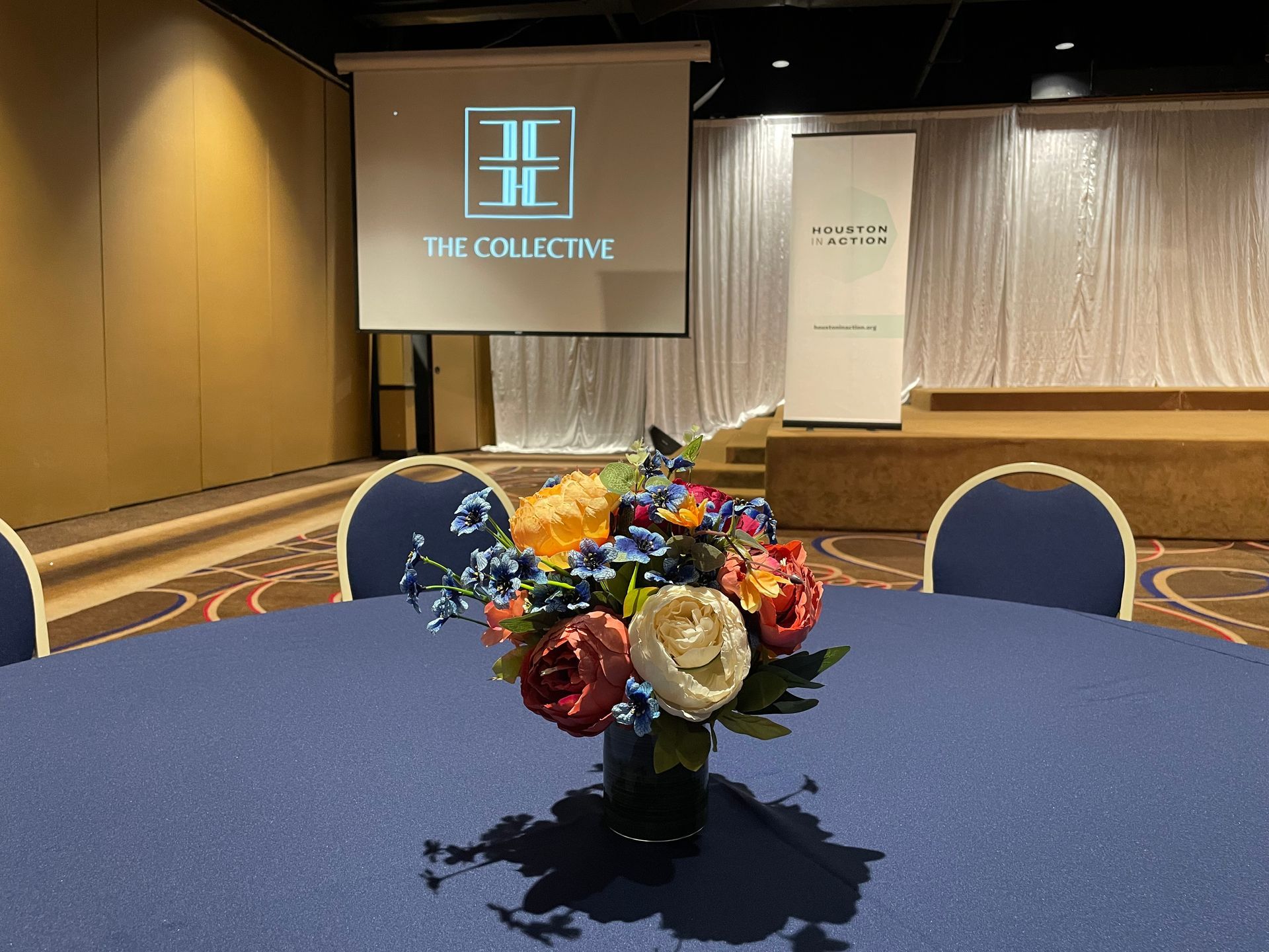 Round table with blue tablecloth, floral centerpiece, in a conference room with a stage, and a screen with a logo.