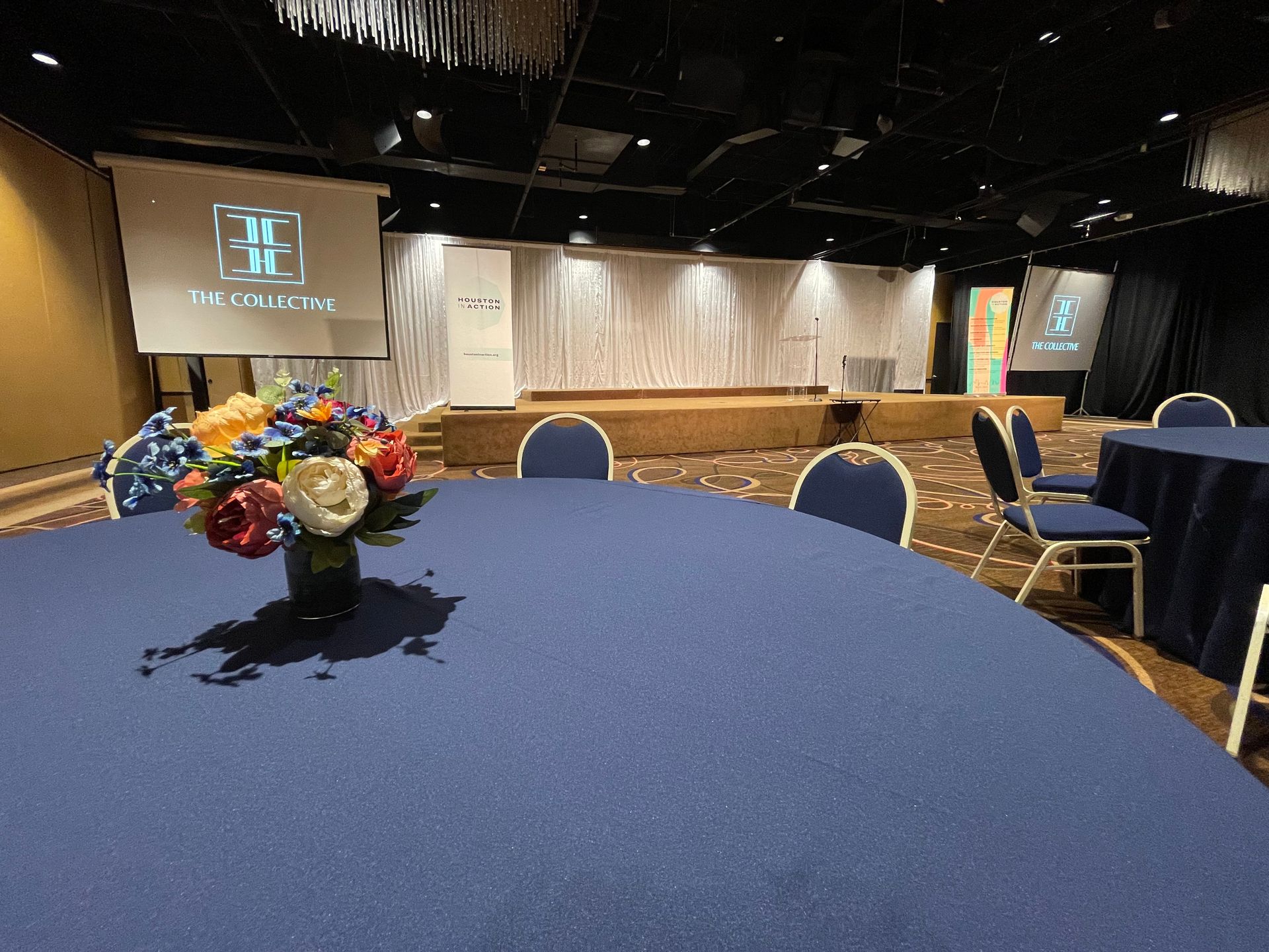 A conference room set up with round tables, a stage, and floral centerpieces.