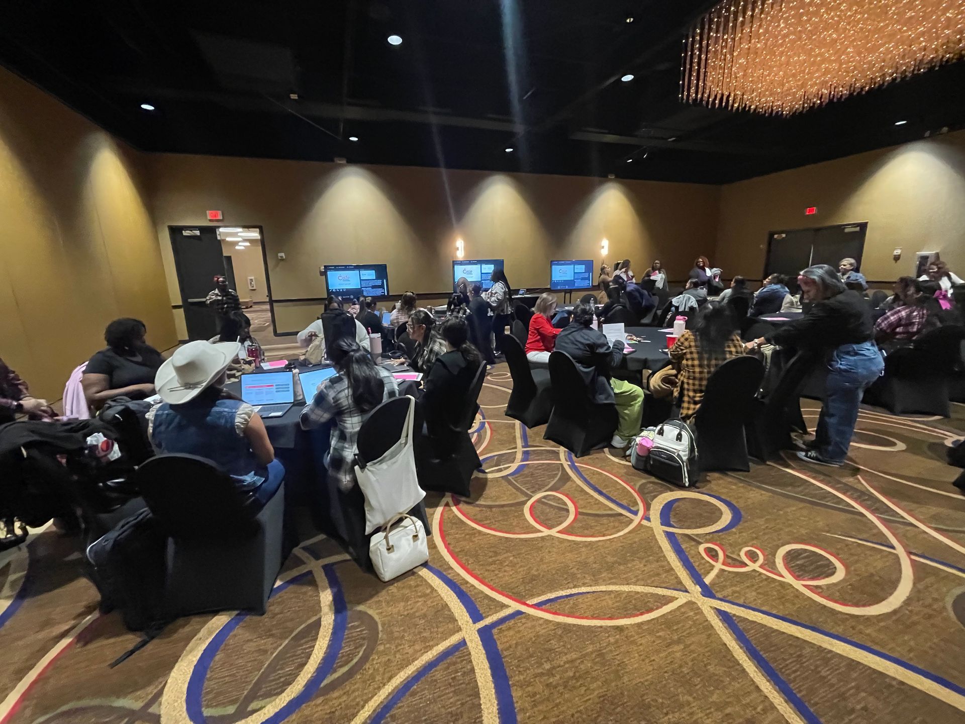 A conference room with people seated at tables, working on laptops. A chandelier hangs from the ceiling.
