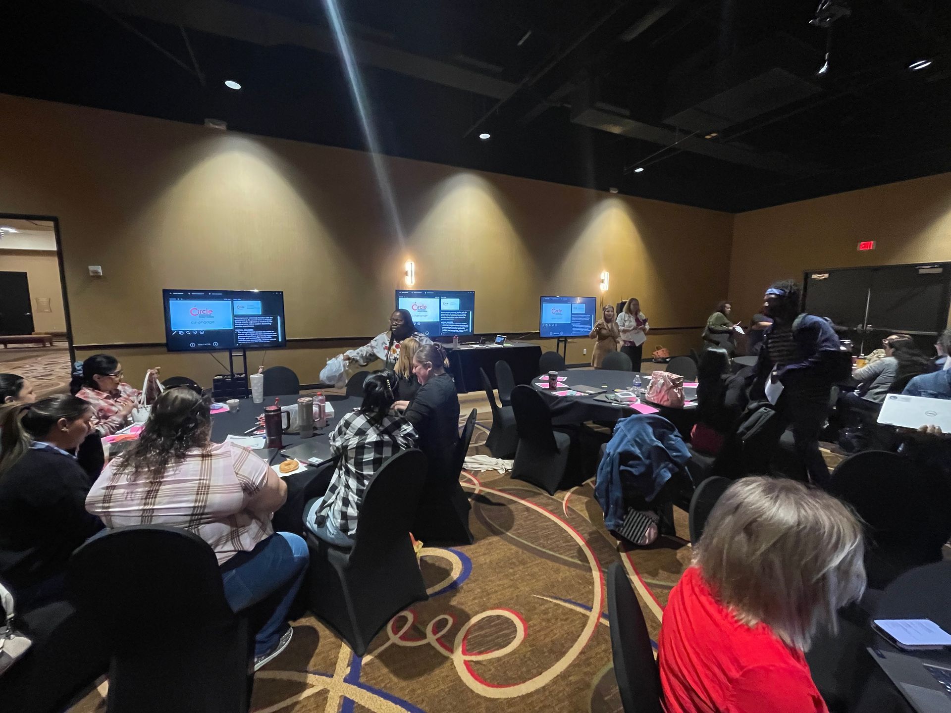 People in a conference room with monitors, seated at tables. Some people are standing.