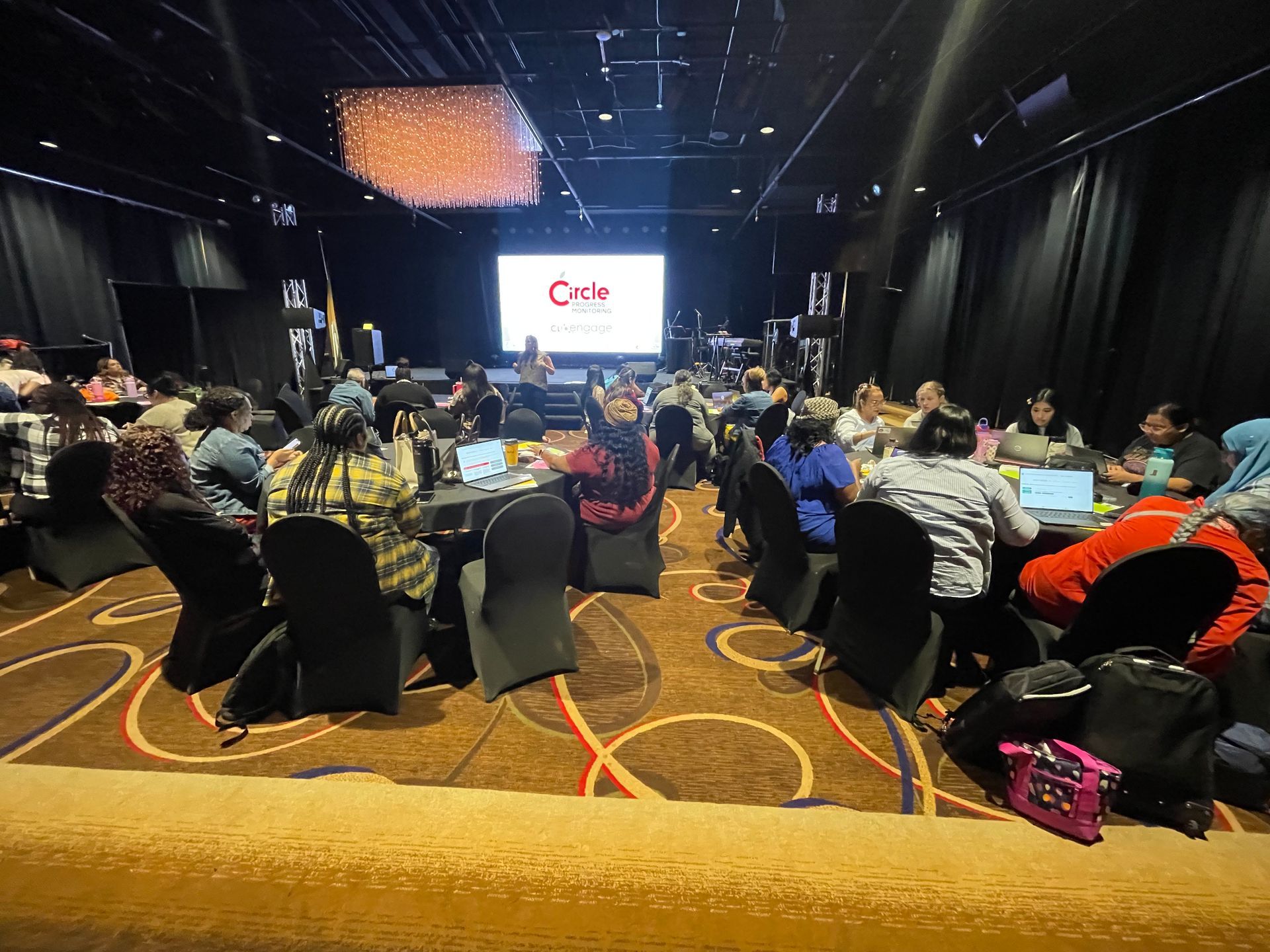 People seated at tables in a conference room, facing a screen with a logo.