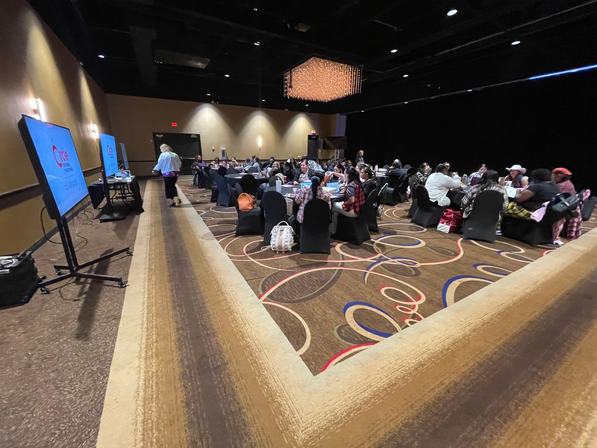 Conference room with attendees seated at tables, two large screens, overhead lighting.