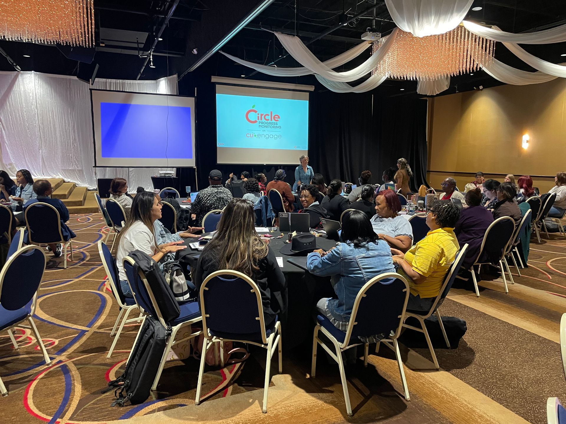 A conference room with a presentation in progress. People seated at round tables. Two screens display images.