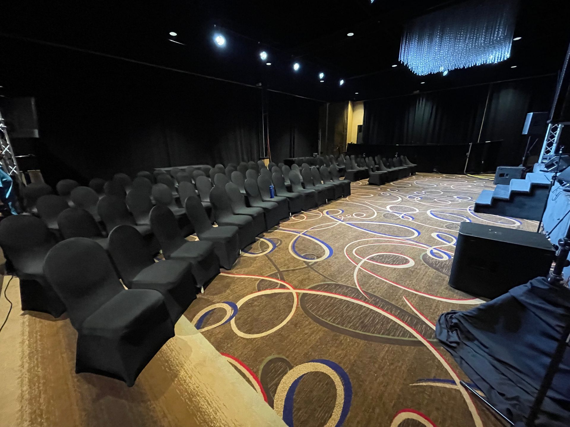 Empty event hall with rows of black chairs, stage, and patterned carpet.