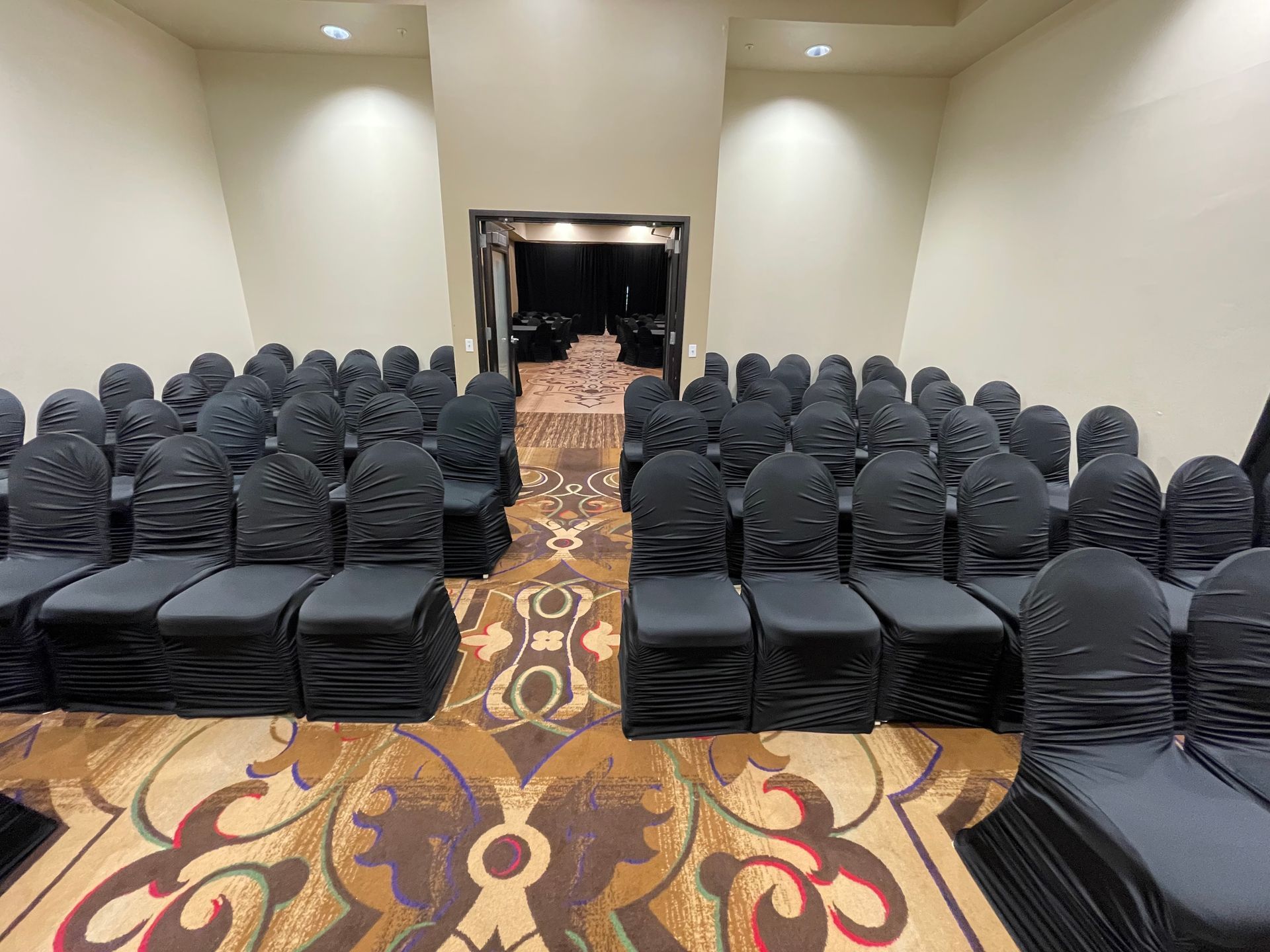 Rows of black chairs face an open doorway in a conference room. Patterned carpet, neutral walls, overhead lights.