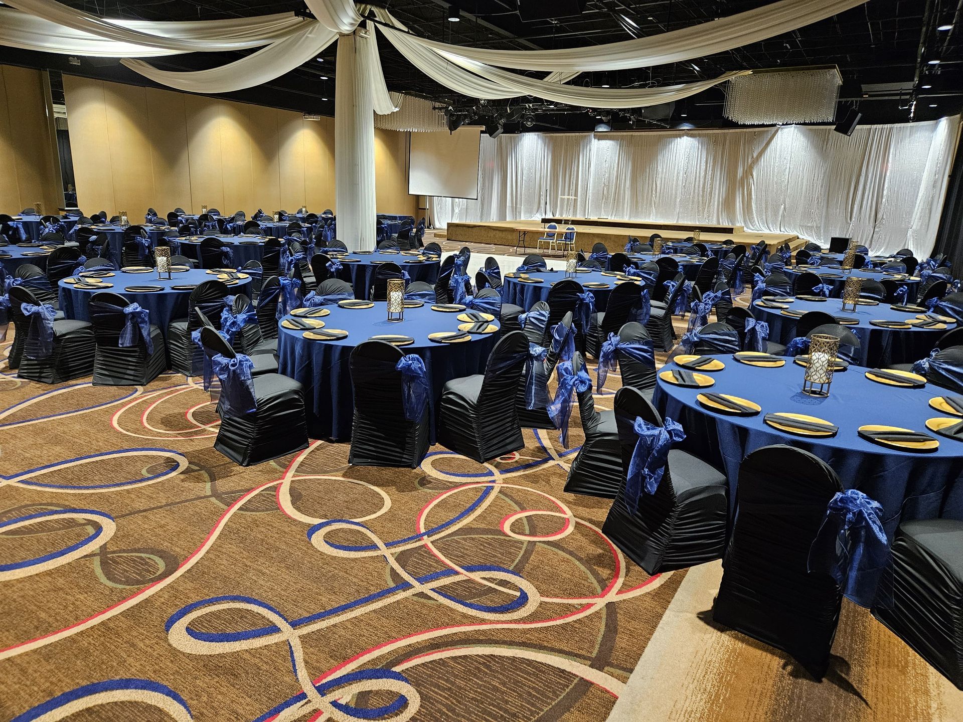 Banquet hall with round tables set with blue linens and chairs with black covers, patterned carpet.