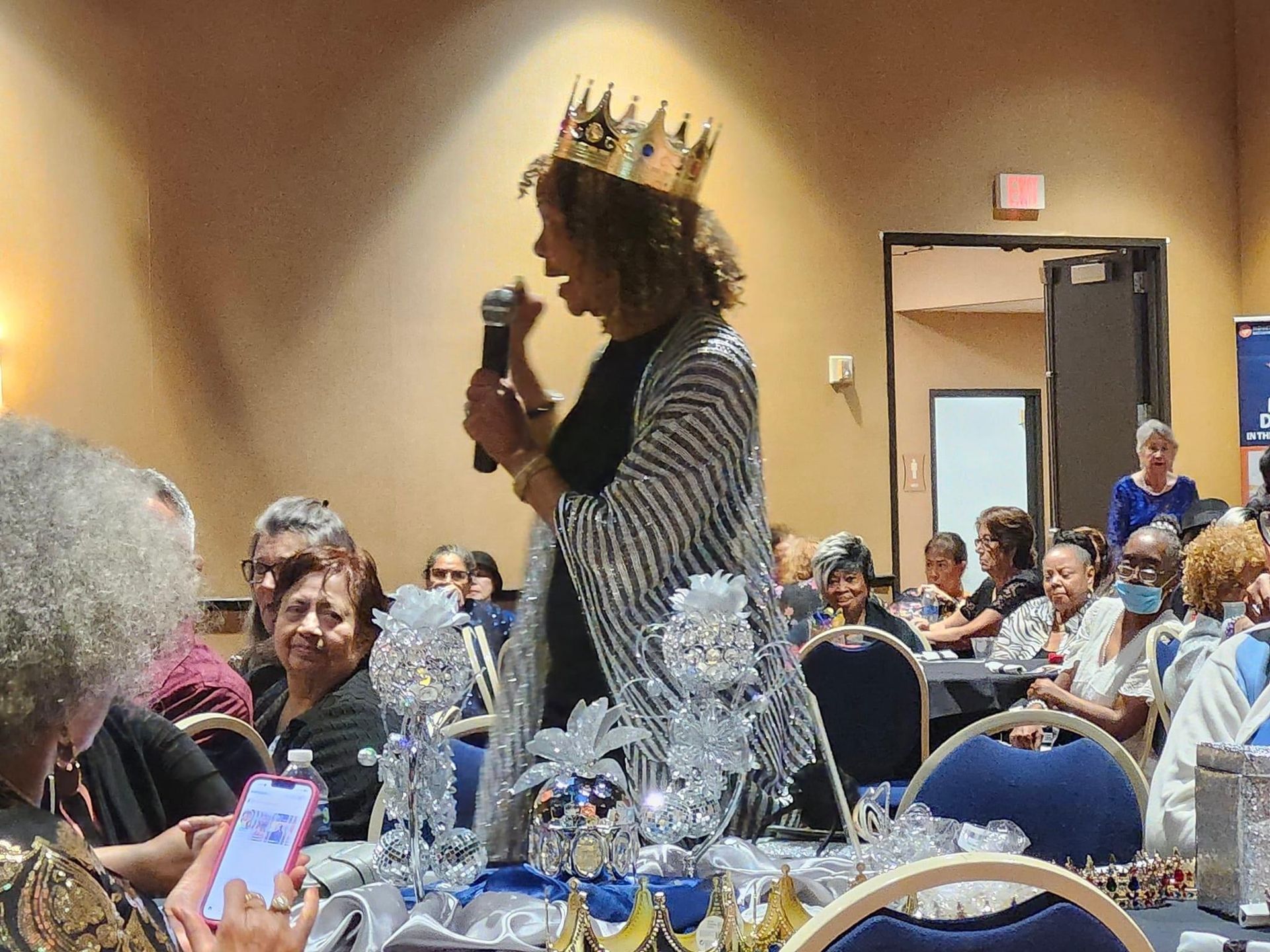 A woman wearing a crown is speaking into a microphone at a senior citizen prom.