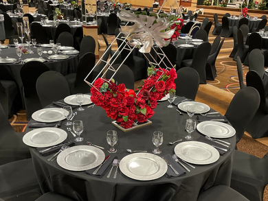 Round banquet table with black chairs, white plates, and red floral centerpiece in a decorated hall