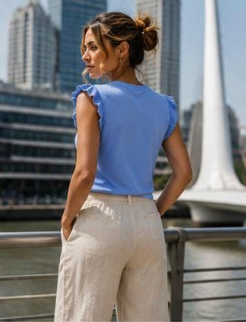 Mujer con blusa azul y pantalones beige de pie junto a una barandilla con el perfil de la ciudad de fondo.