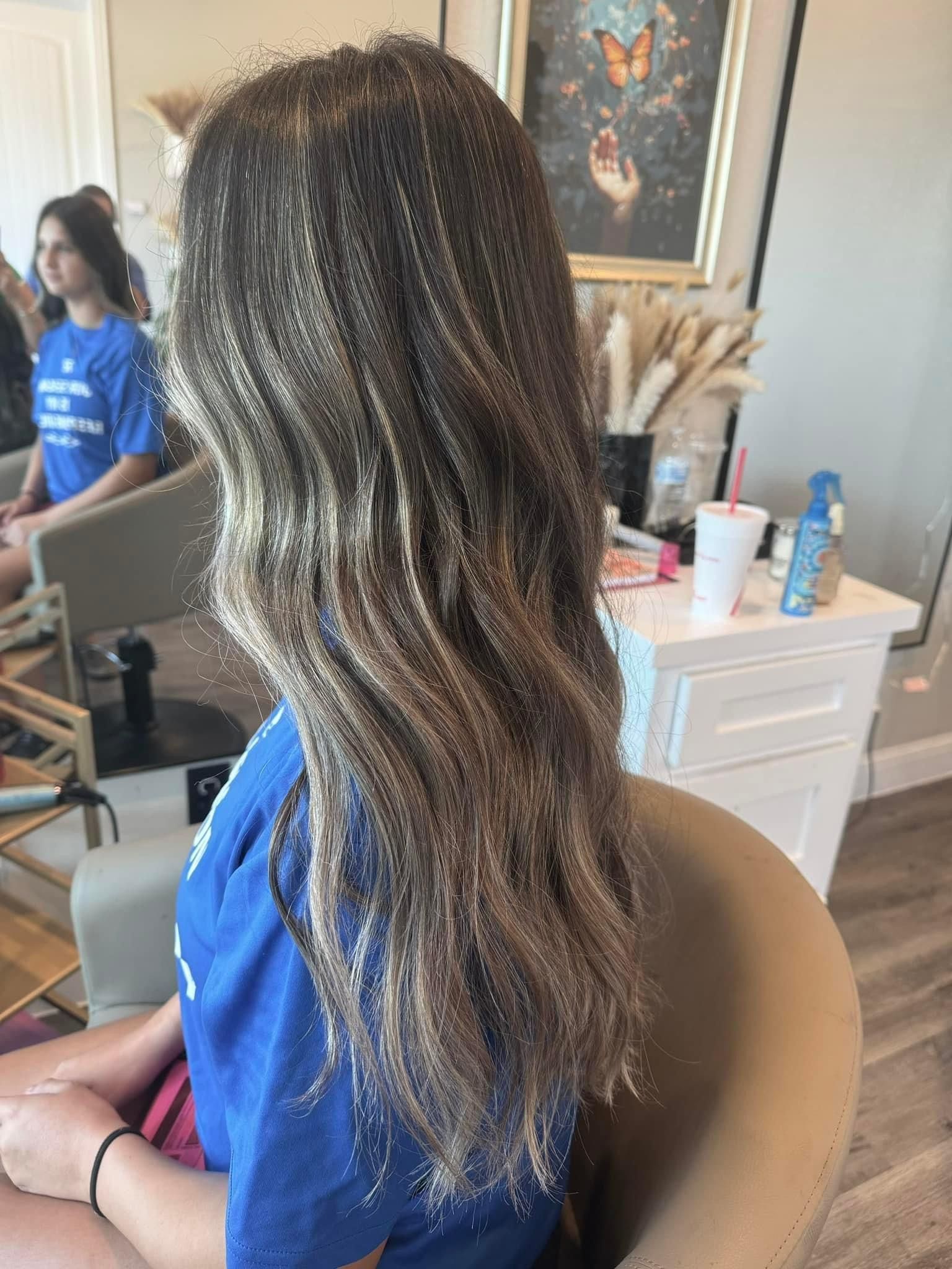 Woman with long wavy hair, highlighted, sitting in salon chair. Another woman in background.