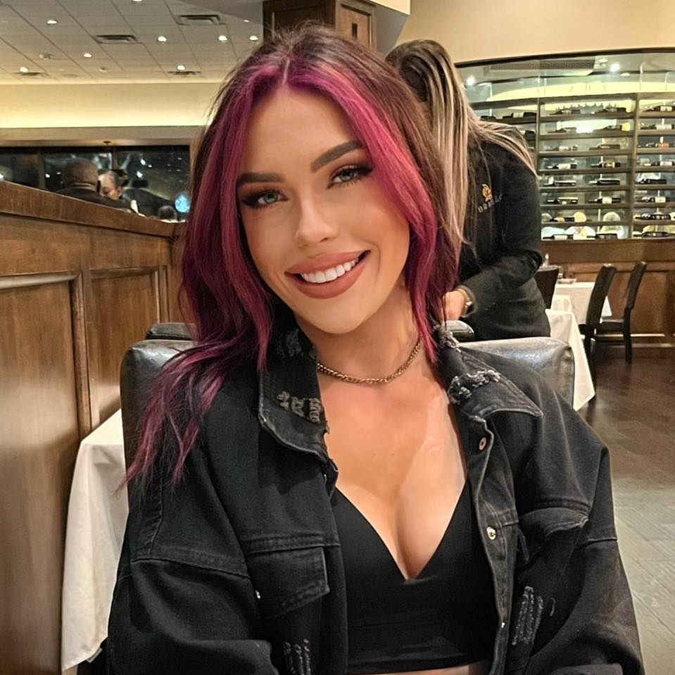 Woman with pink-streaked hair smiles, wearing black jacket and necklace at a restaurant.