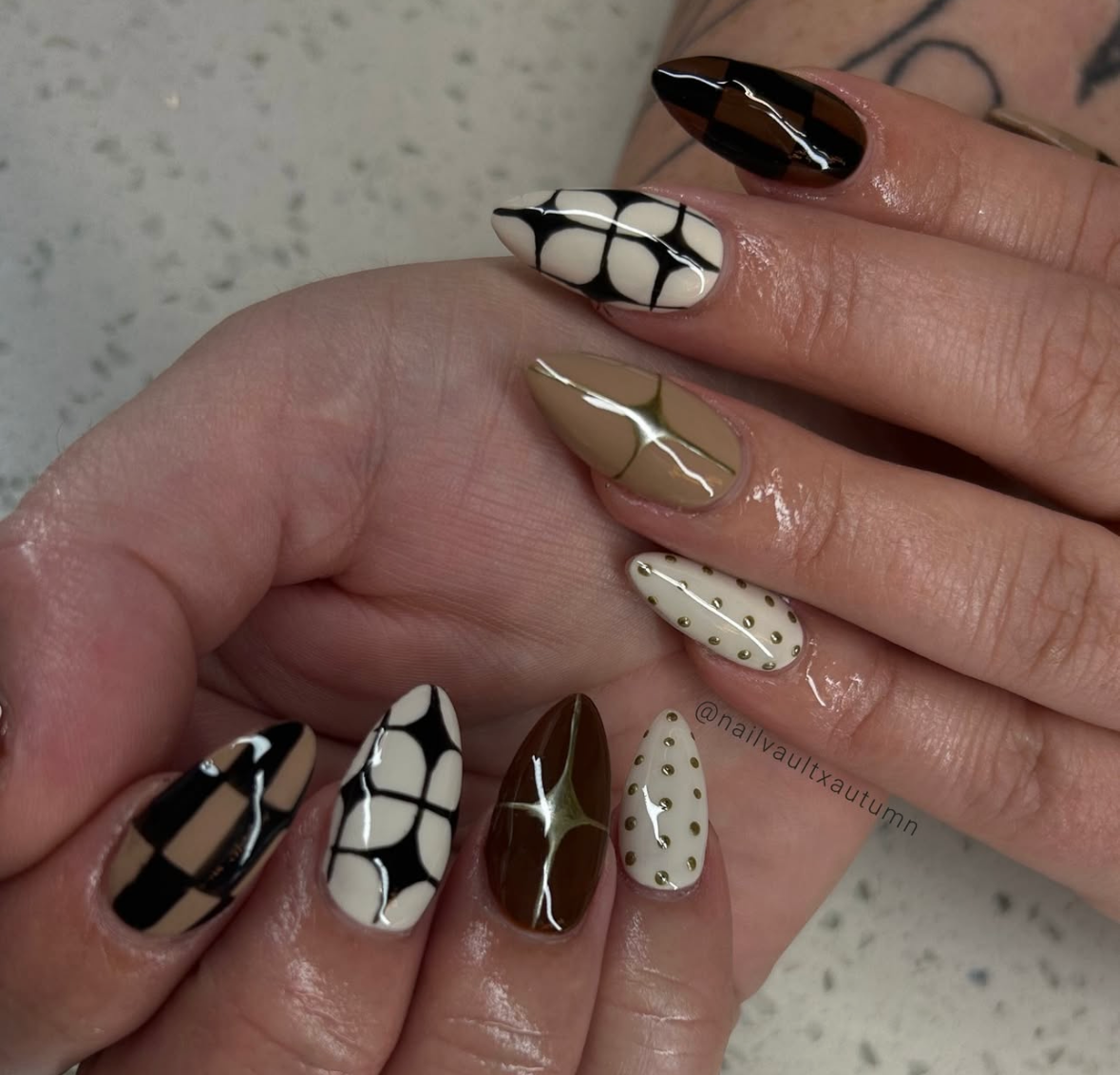 Close-up of a hand with almond-shaped nails decorated in brown, black, and white patterns with a glossy finish.