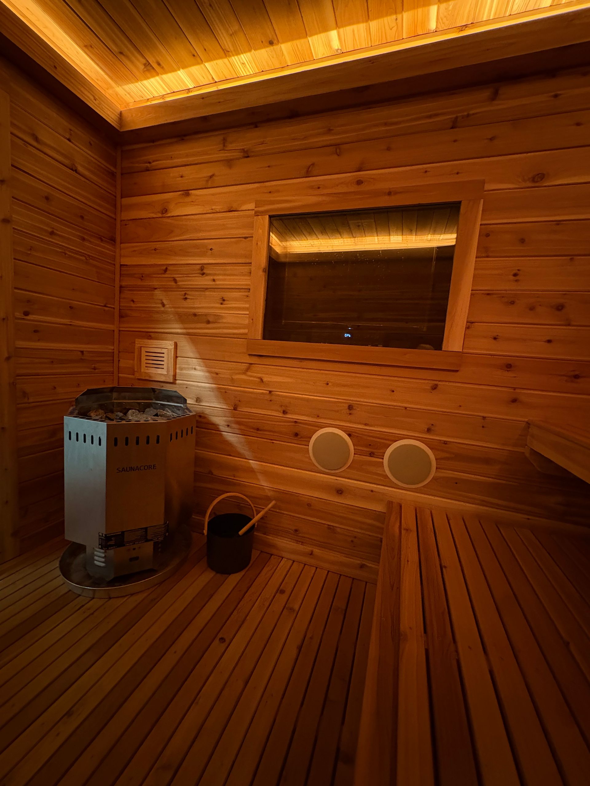 A wooden sauna interior with a heater, bench, and window. Warm light illuminates the space.