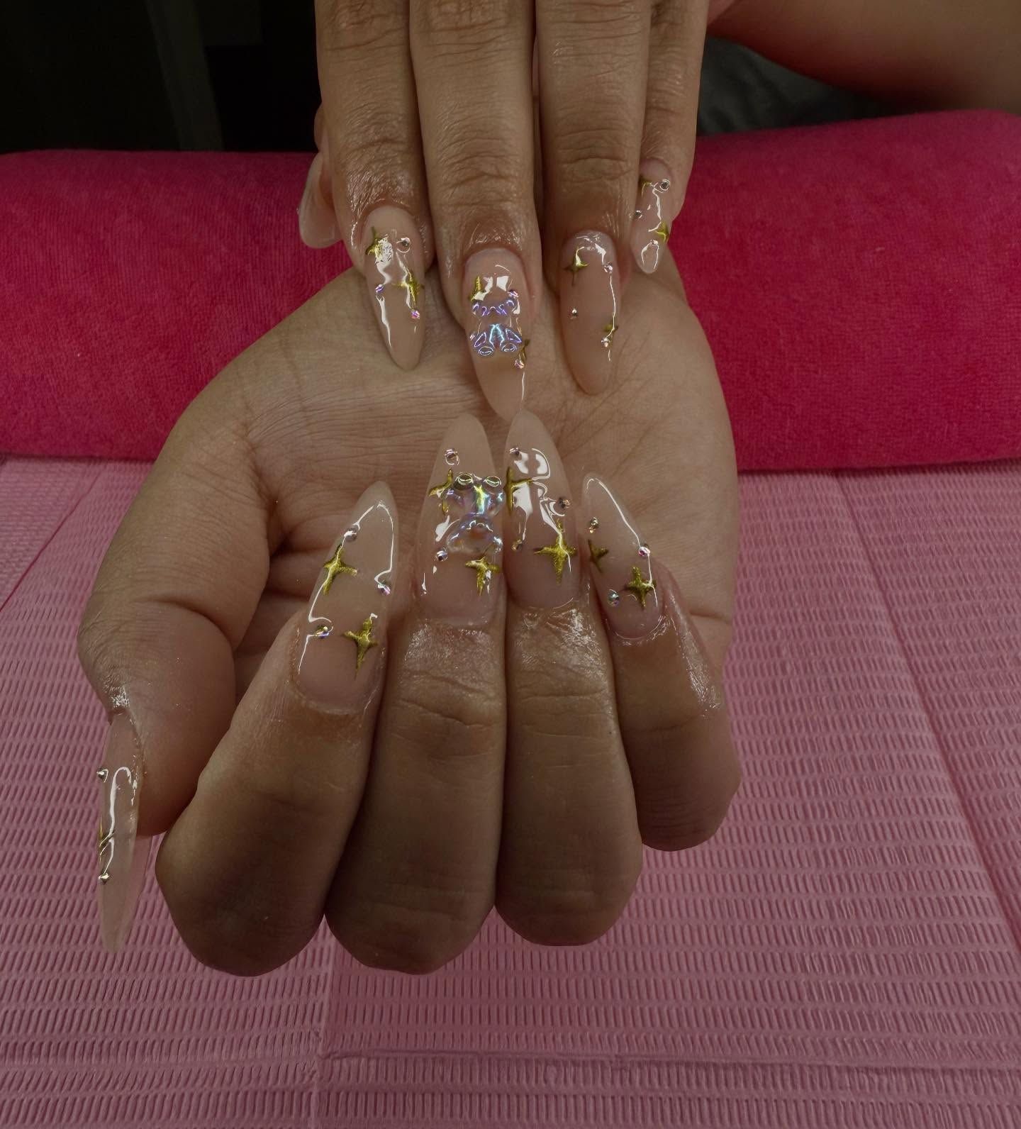 Person's hand with long, clear acrylic nails decorated with gold stars and glitter.