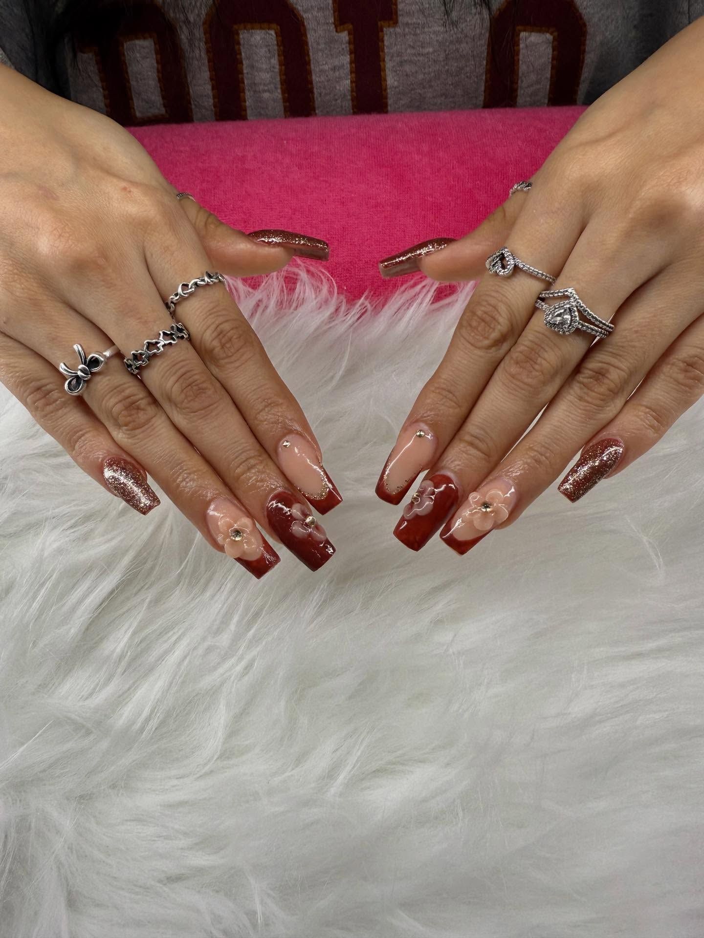 Hands with long, painted nails, featuring a dark red and nude color scheme, adorned with rhinestones, and rings.