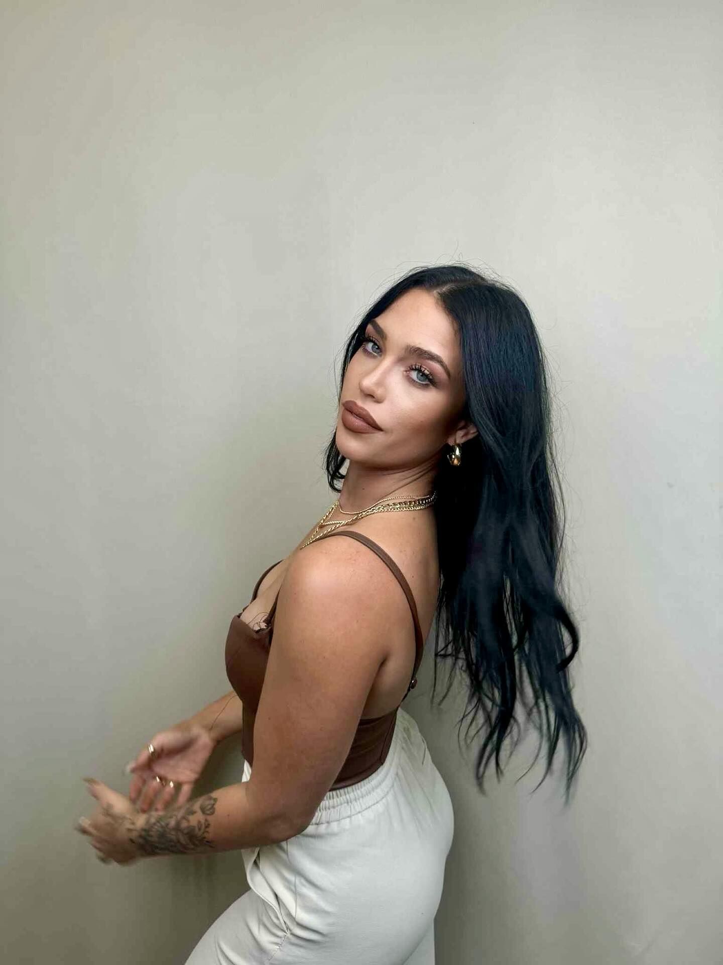 Woman in brown top and white pants, looking over her shoulder, smiling. Gold jewelry and dark hair.