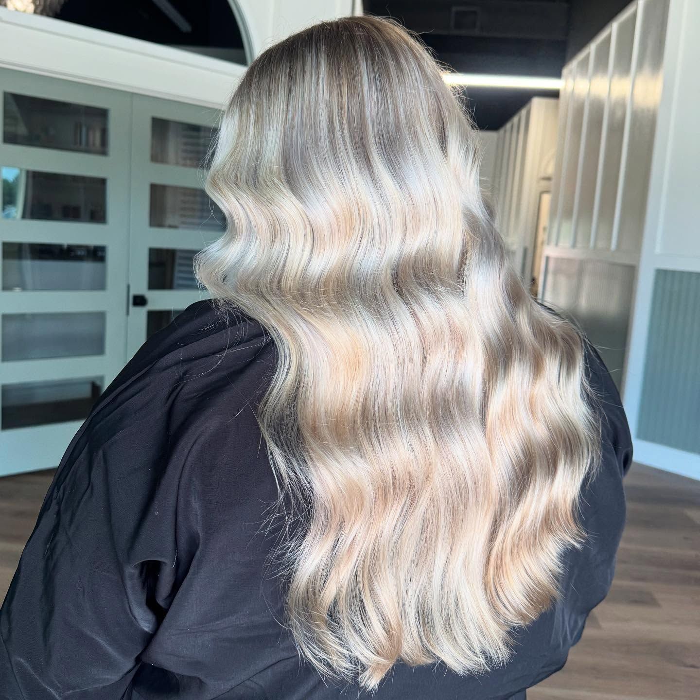 Woman with long, wavy, light blonde hair styled in a salon, wearing a black shirt.