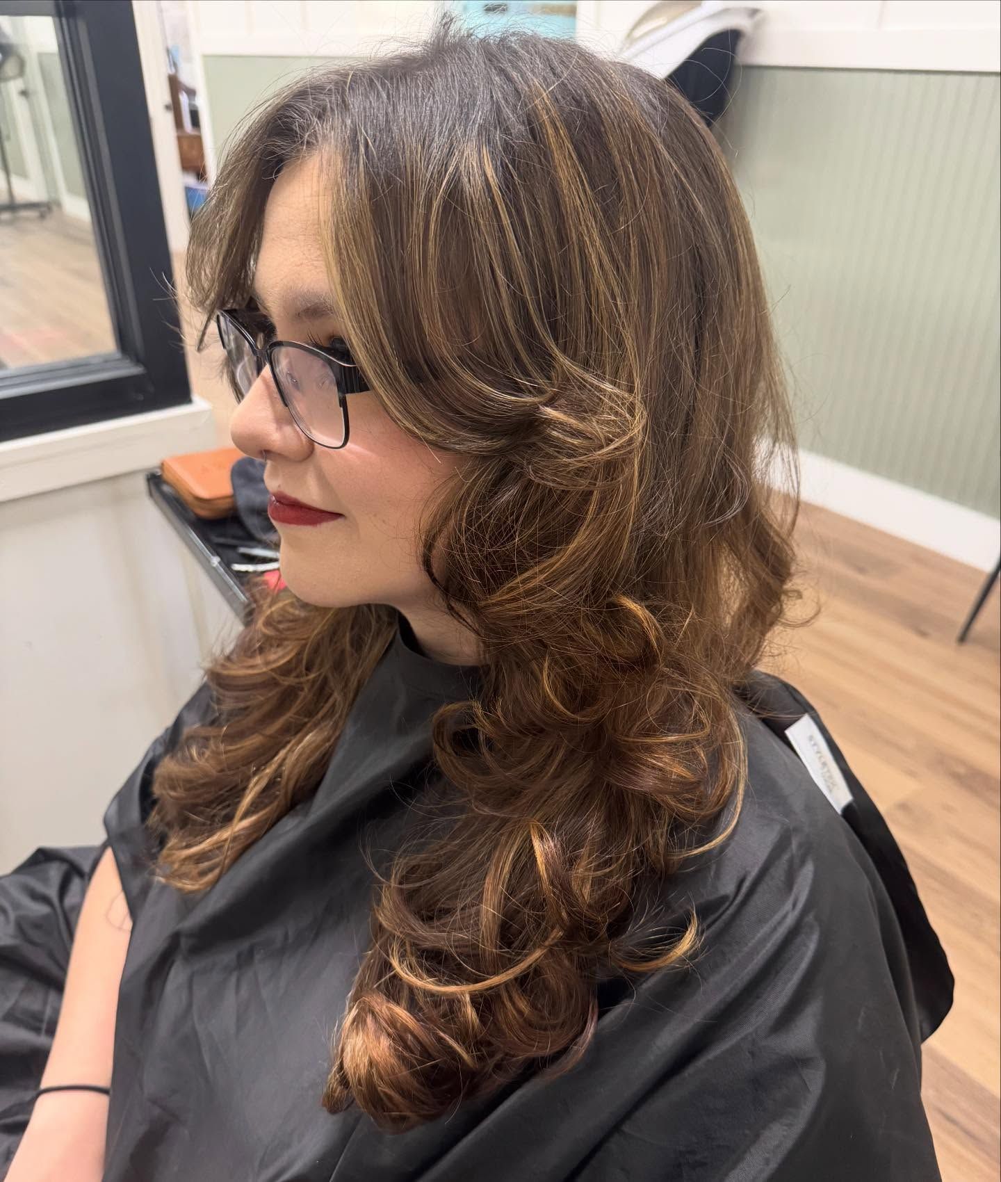 Woman with brown hair, highlights, and layered cut at a salon, wearing glasses.
