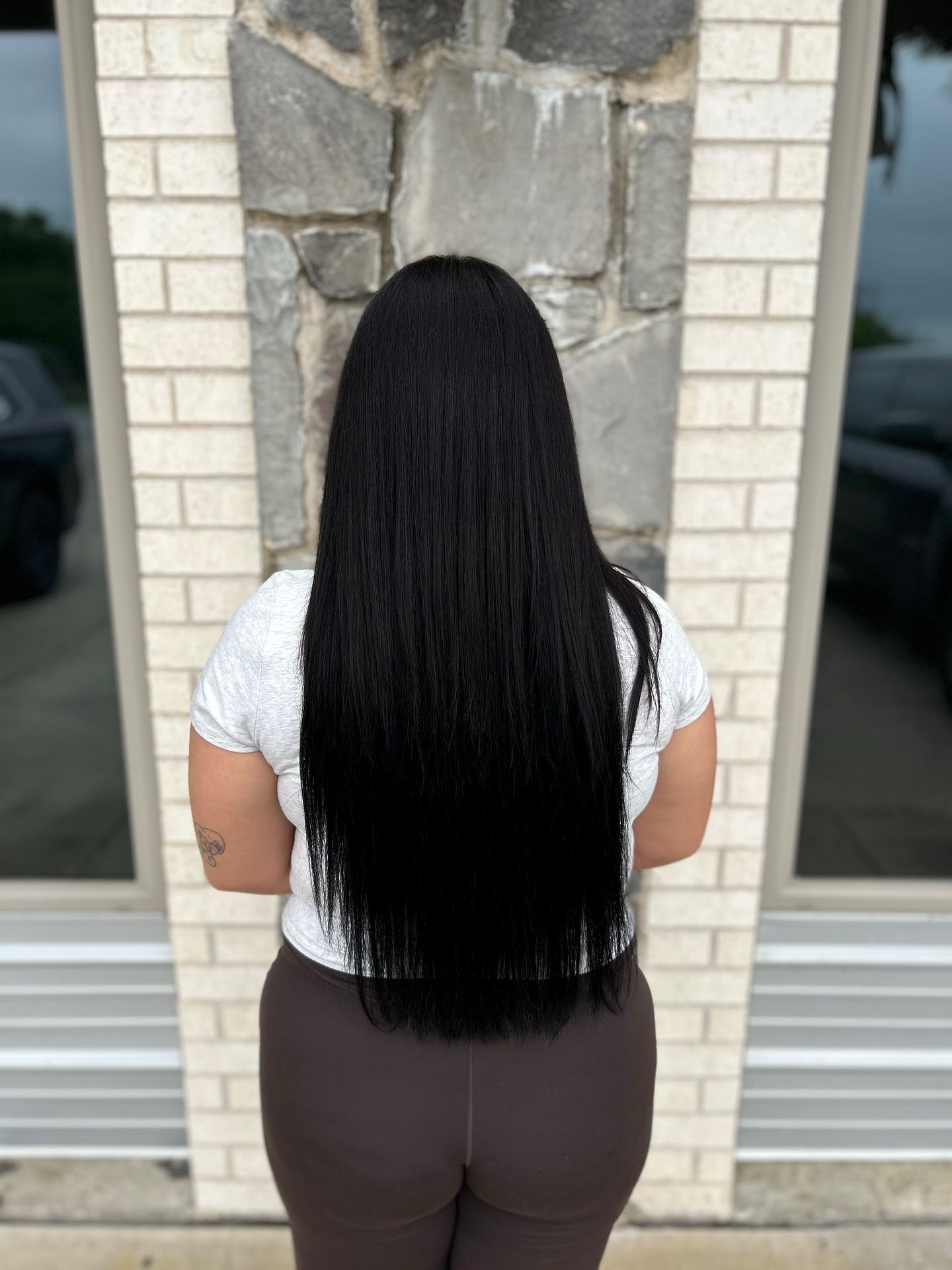 Woman with long, black hair stands outdoors against a stone and brick wall. She wears a white top and brown leggings.