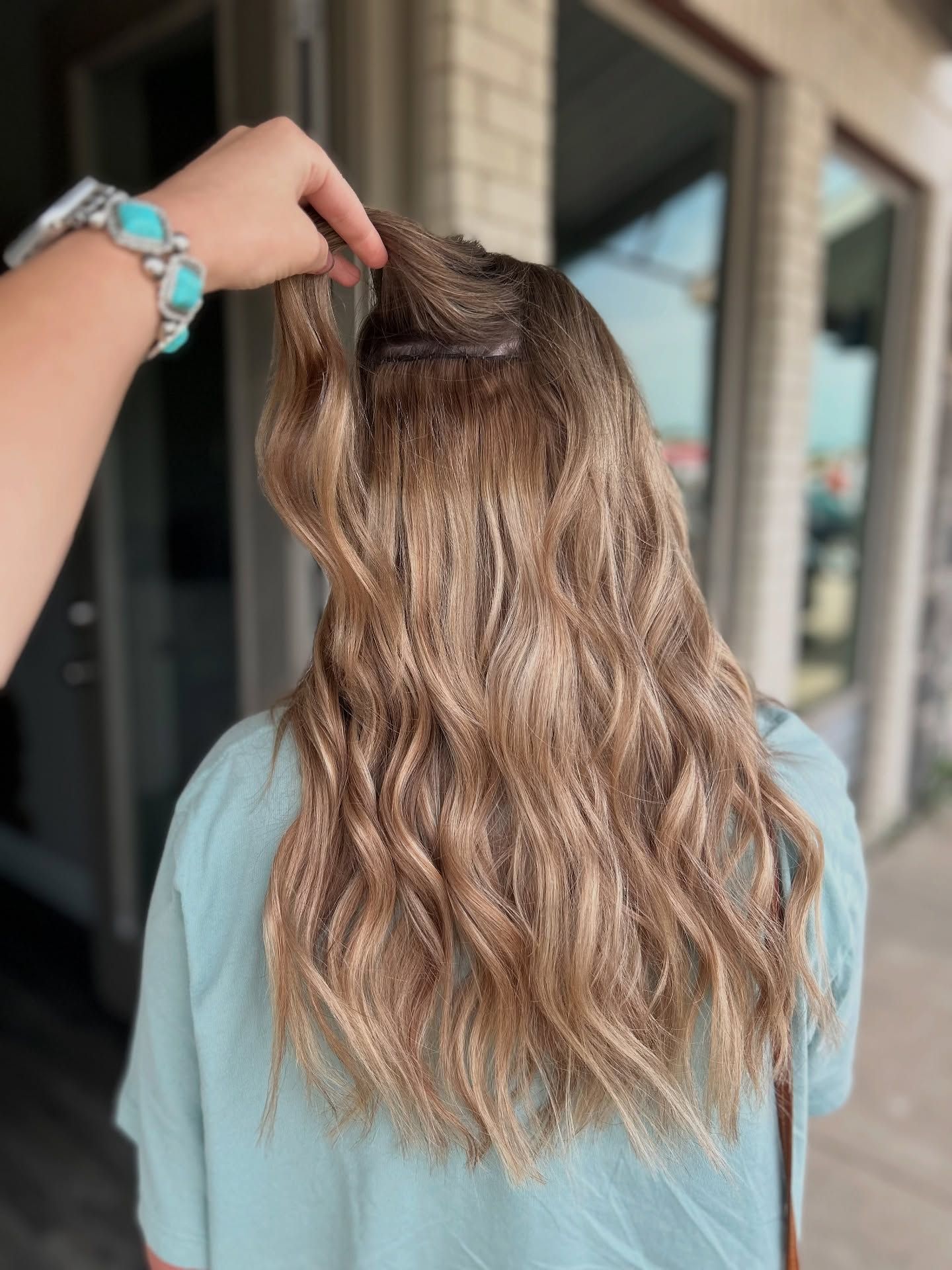 Person's back with long, wavy, light brown hair. Hand holding a section of hair. Outdoors, near windows.