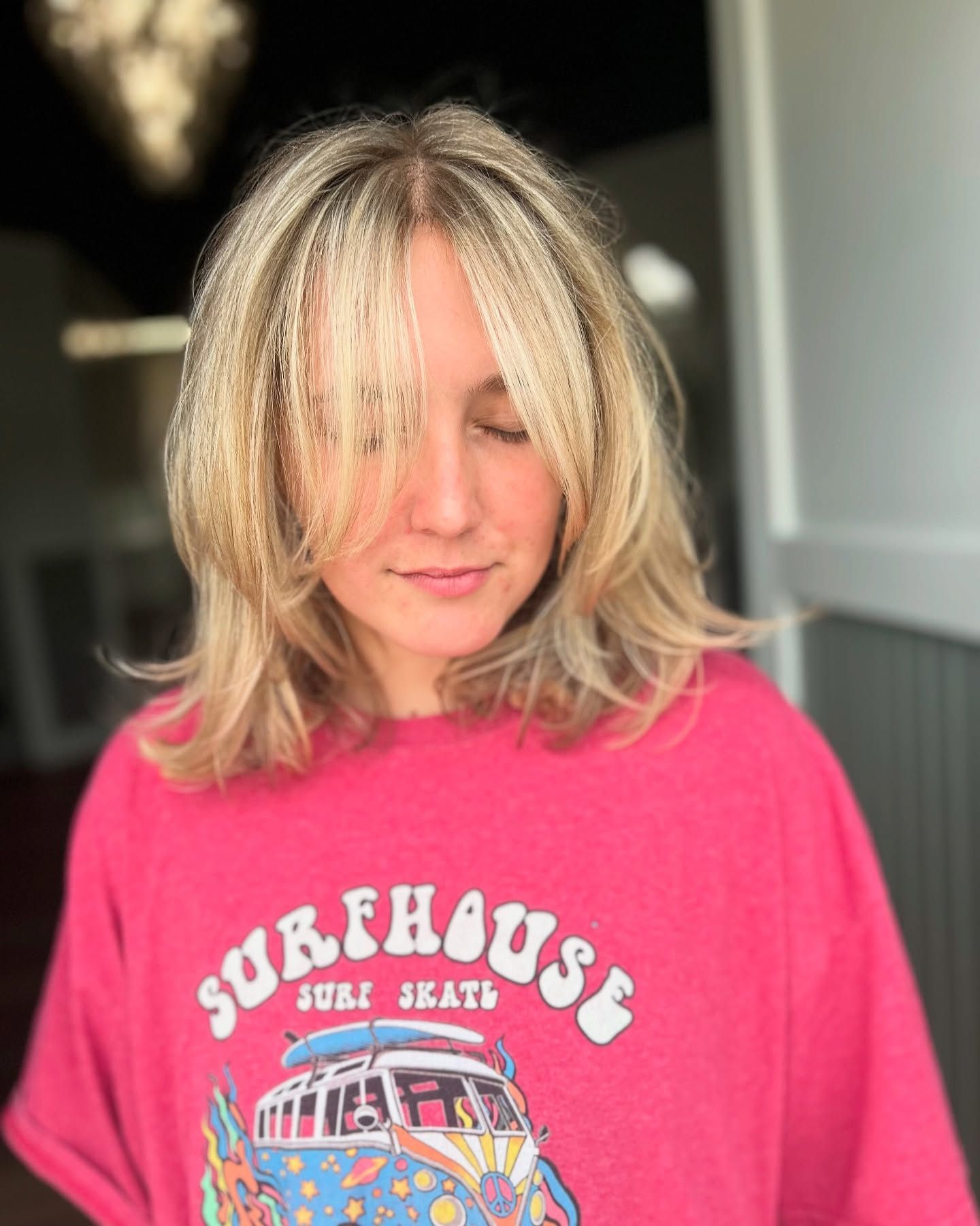 Woman with blonde, layered hair, bangs partially covering her eyes. She wears a pink 