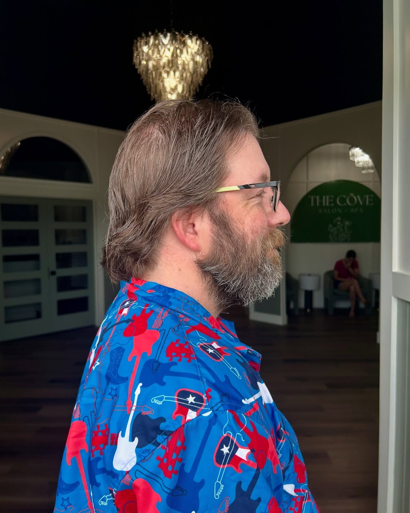 Man in blue shirt with guitar print, glasses, and beard, stands indoors near a door.