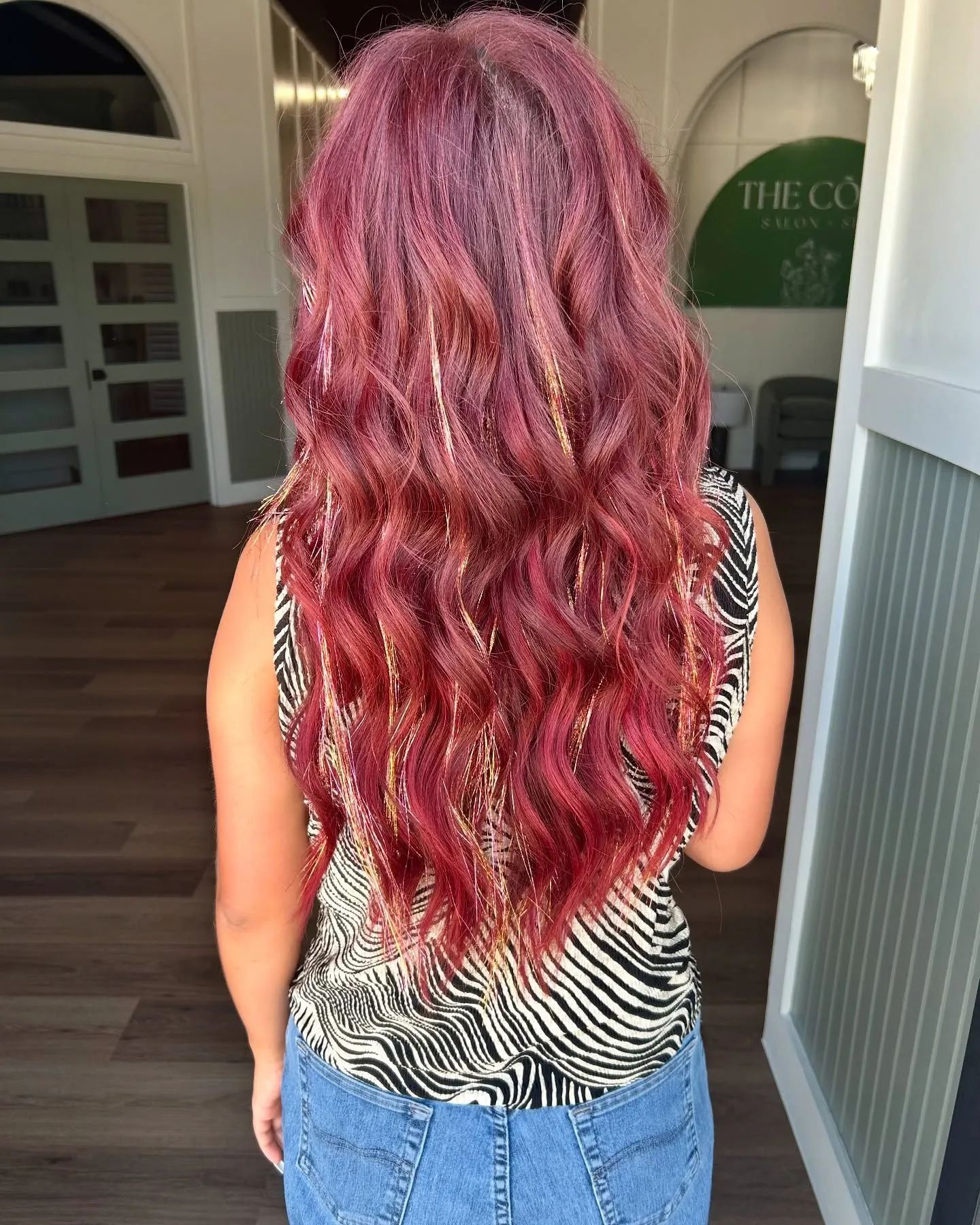 Woman with long wavy magenta hair standing outdoors, seen from behind, wearing a zebra-print top and jeans