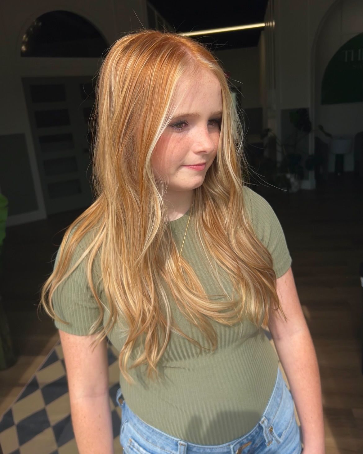 Girl with long, wavy blonde and red hair, wearing a green shirt and denim shorts, indoors.
