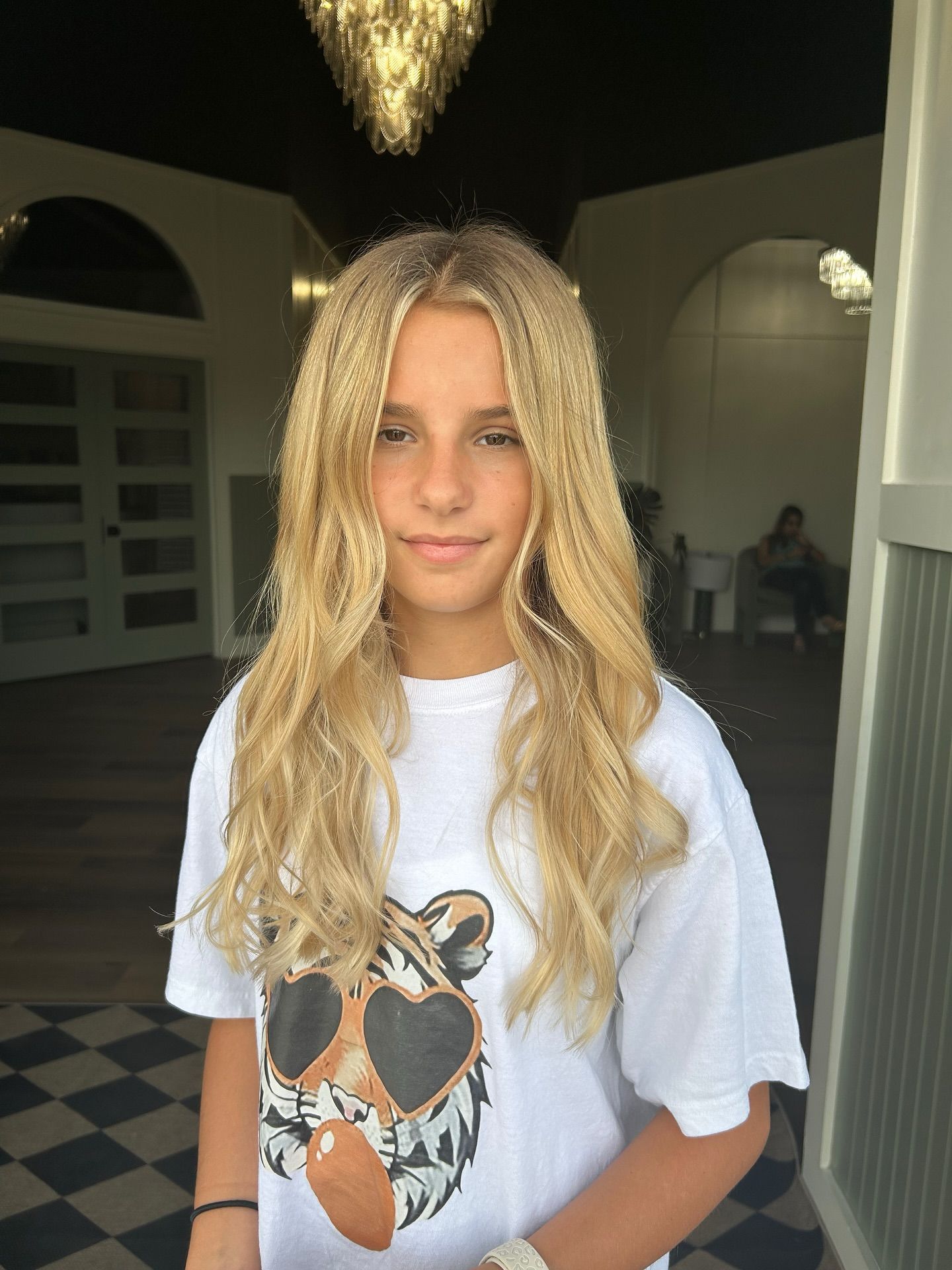 Blonde-haired person with a serious expression, wearing a white t-shirt, standing in a doorway.