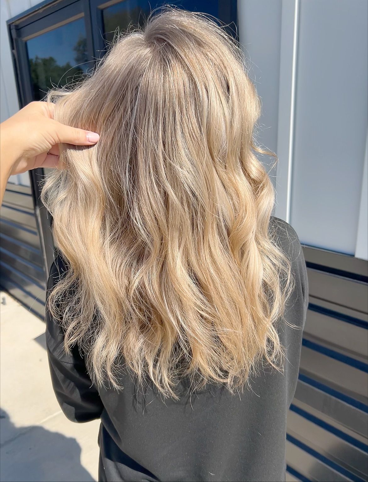 Person with long, wavy, light-colored hair; a hand touches the hair outside by a building.